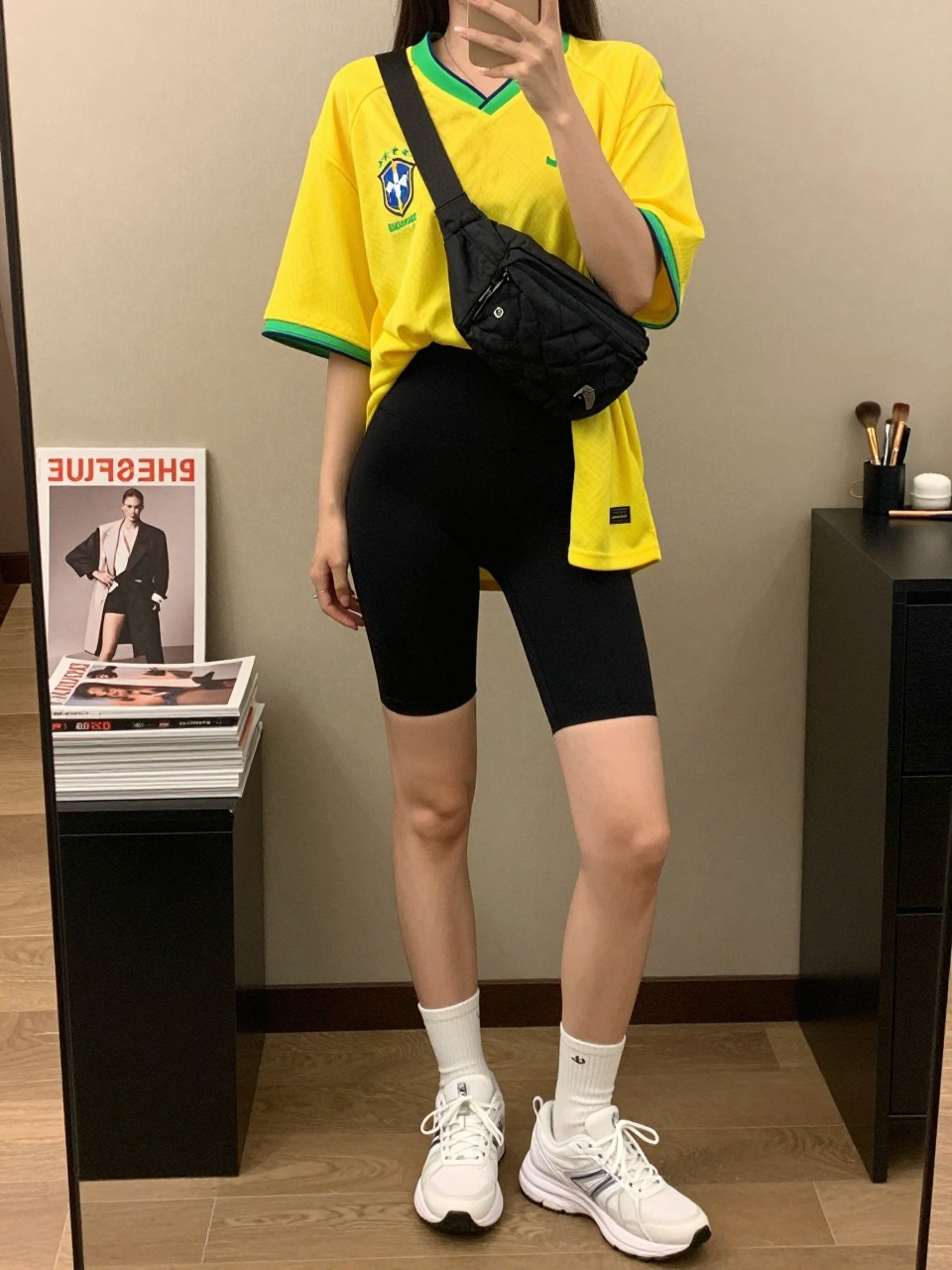 mirror selfie of a woman wearing an oversized yellow Brazil soccer jersey, black high-waisted biker shorts, white athletic sneakers, black nylon belt bag worn crossbody, and white crew socks, clearly showing the same clothing items, colors, fabrics, and accessories listed in the Outfit Pieces section, holding a phone in front of a mirror, cropped at NECK so the face does not show, with a relaxed casual selfie pose, in a fashion studio mirror corner, warm moody lighting, including a stack of fashion magazines on a side table and a makeup brush on the counter, latest iPhone photo quality.