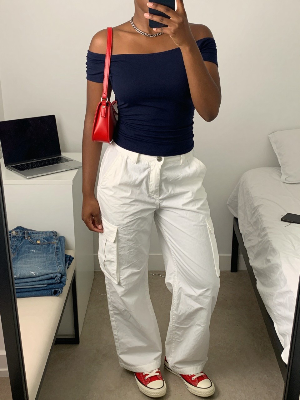 Outfit Image Prompt:
mirror selfie of a Black woman with medium deep brown skin wearing a navy off-shoulder fitted top, white relaxed cargo pants, red low-top sneakers, a silver chain necklace, and a small red crossbody bag, clearly showing the exact outfit pieces listed, holding a phone in front of a mirror, cropped at NECK so the face does not show, relaxed casual selfie pose, in a minimal dressing room, soft indoor lighting, with a laptop on the bed and folded denim jeans on a bench, realistic latest iPhone photo quality, natural skin texture.