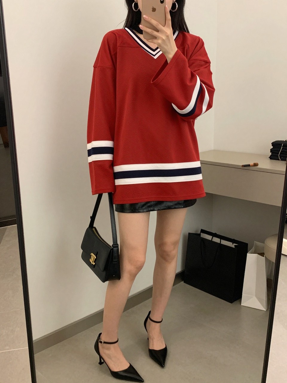 mirror selfie of a woman wearing a red oversized hockey jersey, black faux leather mini skirt, black pointed ankle-strap heels, black mini shoulder bag, and chunky silver hoop earrings, clearly showing the same clothing items, colors, fabrics, and accessories listed in the Outfit Pieces section, holding a phone in front of a mirror, cropped at NECK so the face does not show, with a relaxed casual selfie pose, in a fashion studio mirror corner, soft indoor lighting, including a makeup brush on the counter and shopping bags beside a chair, latest iPhone photo quality.