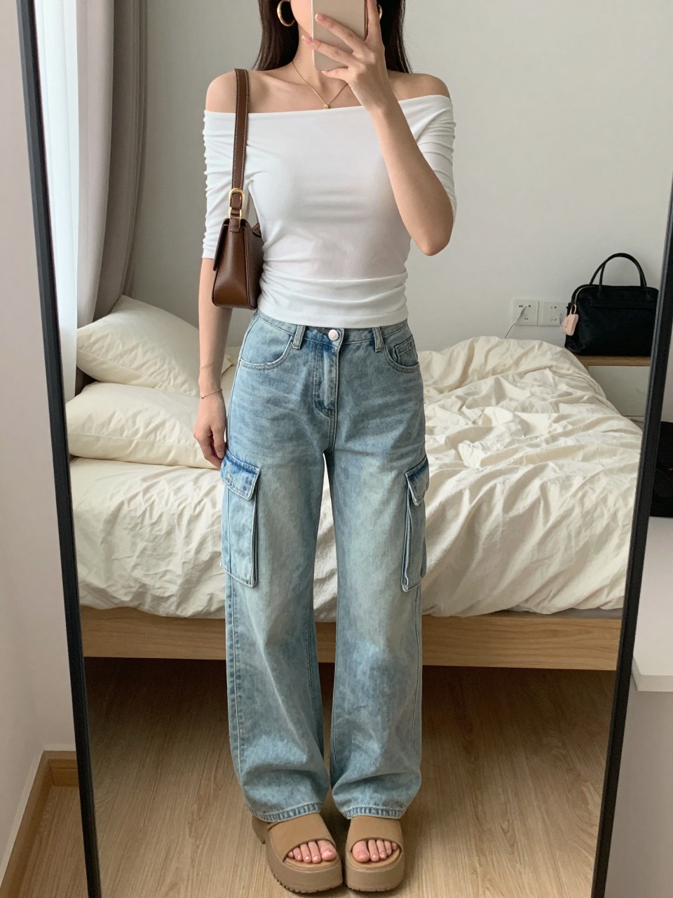 mirror selfie of a woman wearing light wash cargo jeans, fitted white off-shoulder top, tan platform sandals, brown mini shoulder bag, and chunky gold hoop earrings, clearly showing the same clothing items, colors, fabrics, and accessories listed in the Outfit Pieces section, holding a phone in front of a mirror, cropped at NECK so the face does not show, with a relaxed casual selfie pose, in a cozy bedroom mirror setup, natural window light, including soft pillows on an unmade bed and a cosmetic bag on the counter, latest iPhone photo quality.