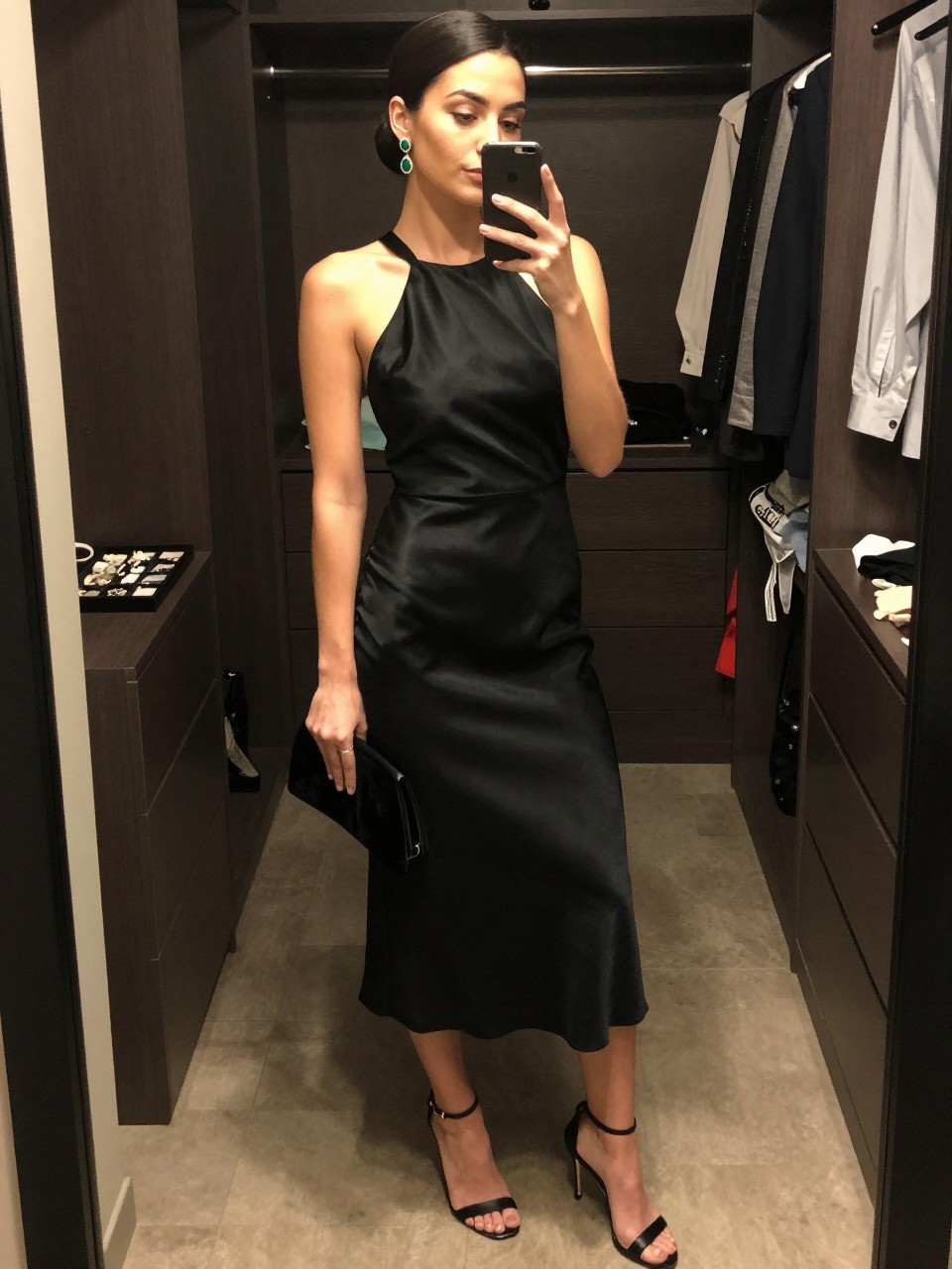 mirror selfie of a woman wearing a black halter neck satin cocktail dress, black stiletto sandals, emerald statement earrings, a black velvet clutch, and a smooth low bun with glowing skin makeup, clearly showing the same clothing items, colors, fabrics, and accessories listed in the Outfit Pieces section, holding a phone in front of a mirror, cropped at NECK so the face does not show, with a relaxed casual selfie pose, in a walk-in closet mirror, warm moody lighting, including open closet with clothes visible and a jewelry tray on the vanity, latest iPhone photo quality.