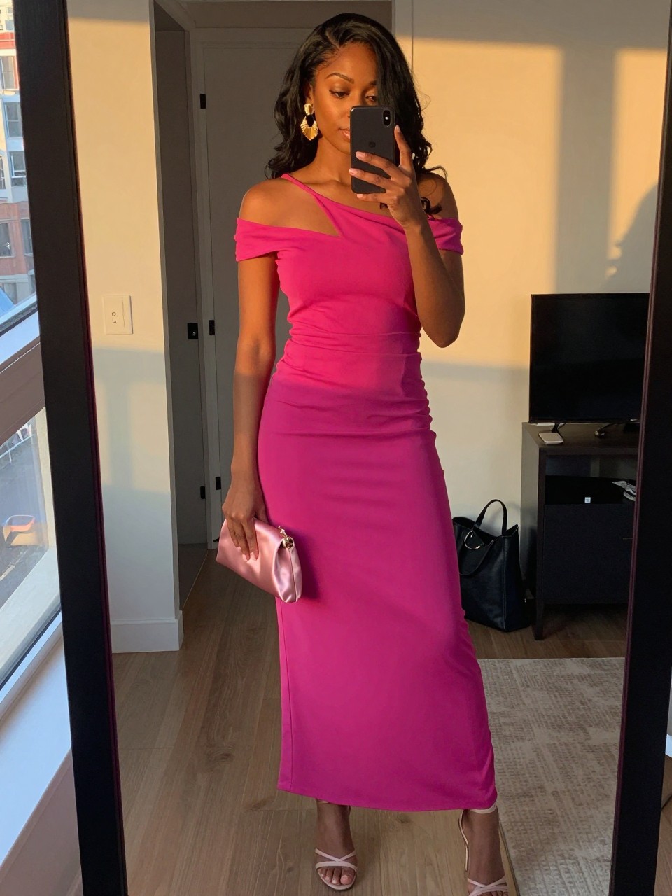 mirror selfie of a black woman wearing a fuchsia off-shoulder maxi dress with nude strappy heels, gold statement earrings, a satin mini clutch, and loose waves hairstyle, holding a phone in front of a mirror, cropped at NECK so face doesn't show, with a natural casual selfie pose, in a city apartment, evening golden hour, including small lived-in details like an open closet behind and a tote bag on the floor, latest iPhone photo quality (no watermarks on images)