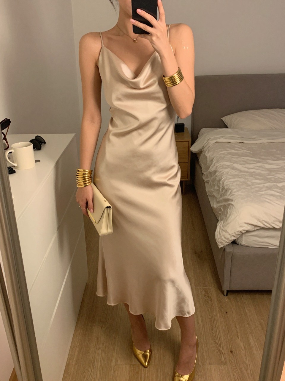 mirror selfie of a woman wearing a champagne satin cowl neck midi dress with gold metallic heels, lightly stacked gold bangles, a cream small clutch, and glowy skin makeup, holding a phone in front of a mirror, cropped at NECK so face doesn't show, with a natural casual selfie pose, in a feminine-themed bedroom, warm moody lighting, including small lived-in details like a coffee mug nearby and sunglasses on a dresser, latest iPhone photo quality (no watermarks on images)