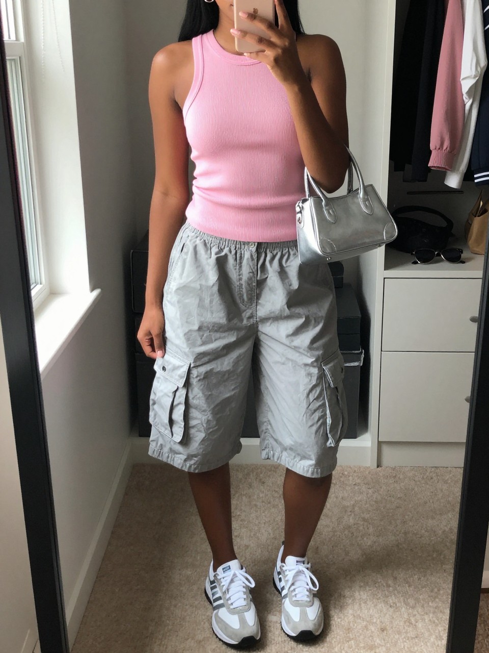 mirror selfie of a Black woman wearing gray baggy cargo shorts, baby pink fitted knit tank top, white and silver sneakers, silver mini handbag, and small silver huggie earrings, clearly showing the same clothing items, colors, fabrics, and accessories listed in the Outfit Pieces section, holding a phone in front of a mirror, cropped at NECK so the face does not show, with a relaxed casual selfie pose, in a walk-in closet mirror, natural window light, including an open closet with clothes visible and sunglasses placed on a dresser, latest iPhone photo quality.