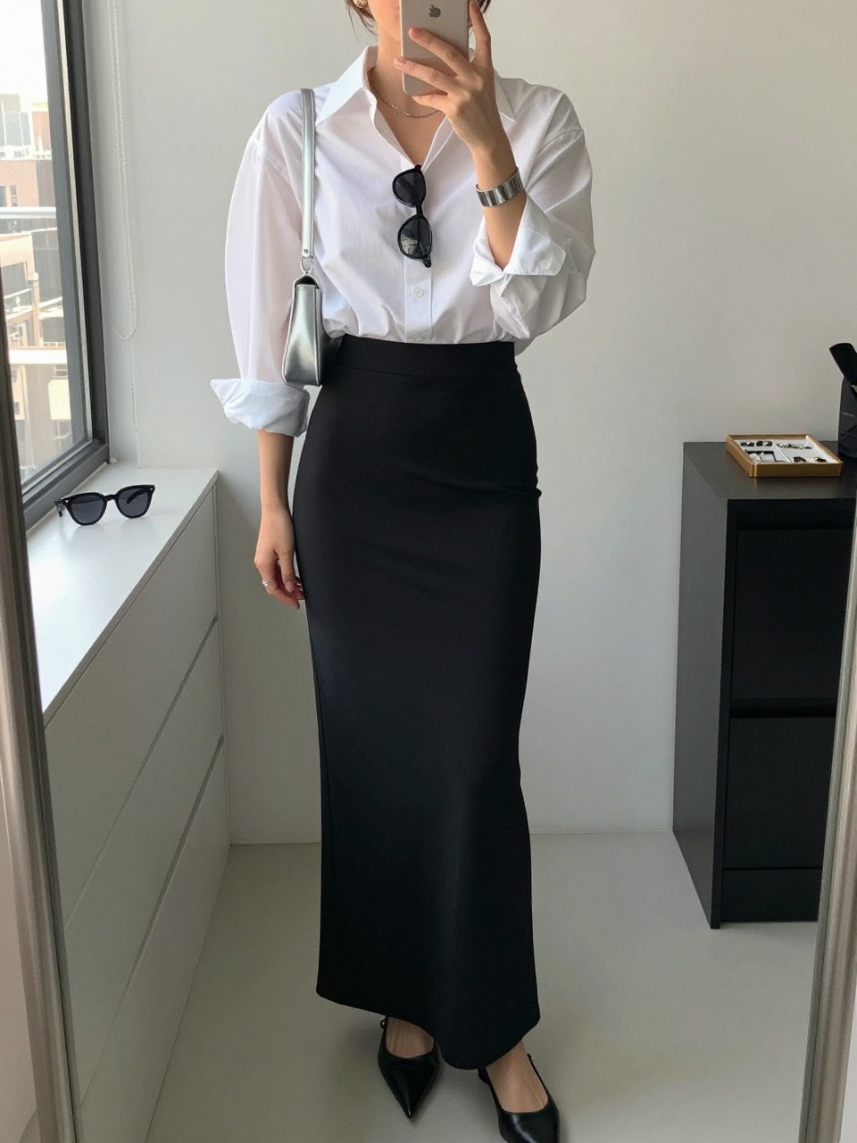 Outfit Image Prompt:
mirror selfie of a woman wearing a crisp oversized white poplin button-down shirt, black fitted maxi column skirt, black pointed slingback flats, slim black sunglasses, small silver shoulder bag, and silver cuff bracelet, holding a phone in front of a mirror, cropped at NECK so the face does not show, relaxed NYC mirror selfie pose, in a minimal dressing room, natural window light, with sunglasses placed on a dresser and a jewelry tray on the vanity, realistic latest iPhone photo quality, clean high-fashion styling.