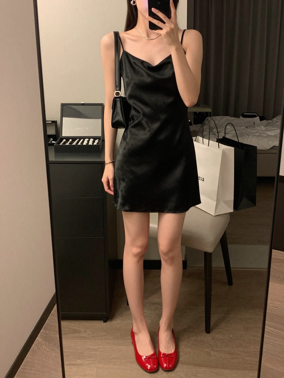 mirror selfie of a woman wearing a black satin slip dress, glossy red jelly shoes, black mini shoulder bag, silver hoop earrings, and a slim silver necklace, clearly showing the same clothing items, colors, fabrics, and accessories listed in the Outfit Pieces section, holding a phone in front of a mirror, cropped at NECK so the face does not show, with a relaxed casual selfie pose, in a stylish city apartment, warm moody lighting, including a jewelry tray on the vanity and shopping bags beside a chair, latest iPhone photo quality.