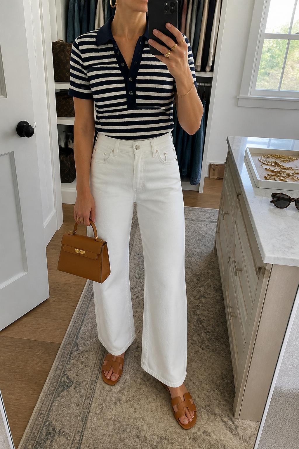 mirror selfie of a woman wearing a navy and white striped short-sleeve knit polo shirt tucked into white high-waisted wide-leg jeans, tan leather slide sandals, tan structured mini bag, and gold hoop earrings, clearly showing the same clothing items, colors, fabrics, and accessories listed in the Outfit Pieces section, holding a phone in front of a mirror, cropped at NECK so the face does not show, with a relaxed casual selfie pose, in a walk-in closet mirror, natural window light, including sunglasses placed on a dresser and a jewelry tray on the vanity, latest iPhone photo quality.
