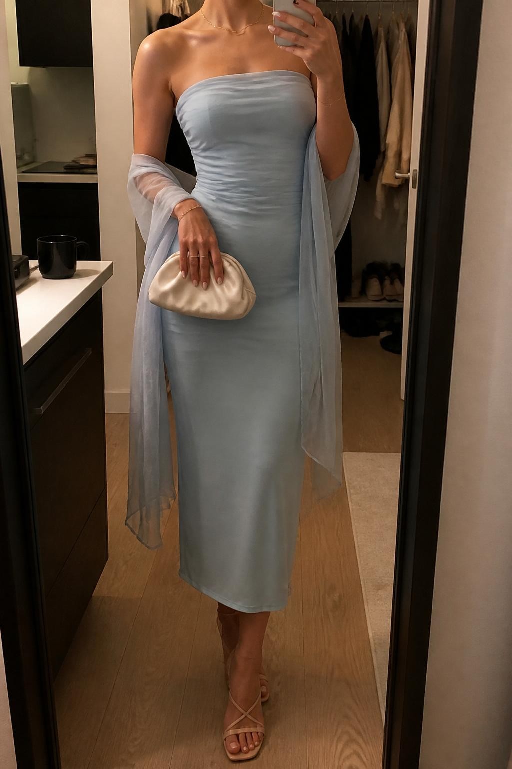 mirror selfie of a woman wearing a pale blue strapless midi dress with a matching sheer wrap, nude strappy heels, a satin clutch bag, and glossy lip tint, holding a phone in front of a mirror, cropped at NECK so face doesn't show, with a natural casual selfie pose, in a city apartment, warm moody lighting, including small lived-in details like a coffee mug nearby and an open closet behind, latest iPhone photo quality (no watermarks on images)