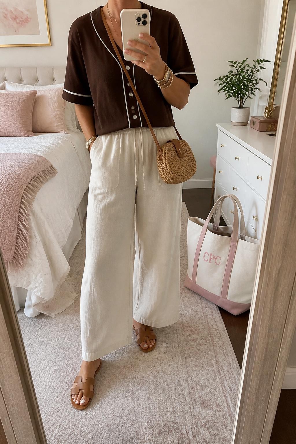 mirror selfie of a woman wearing a brown button-front baseball jersey, cream high-waisted linen pants, tan leather slide sandals, woven straw crossbody bag, and gold layered bracelets, clearly showing the same clothing items, colors, fabrics, and accessories listed in the Outfit Pieces section, holding a phone in front of a mirror, cropped at NECK so the face does not show, with a relaxed casual selfie pose, in a feminine-themed bedroom, soft indoor lighting, including a small plant on a dresser and a tote bag resting on the floor, latest iPhone photo quality.