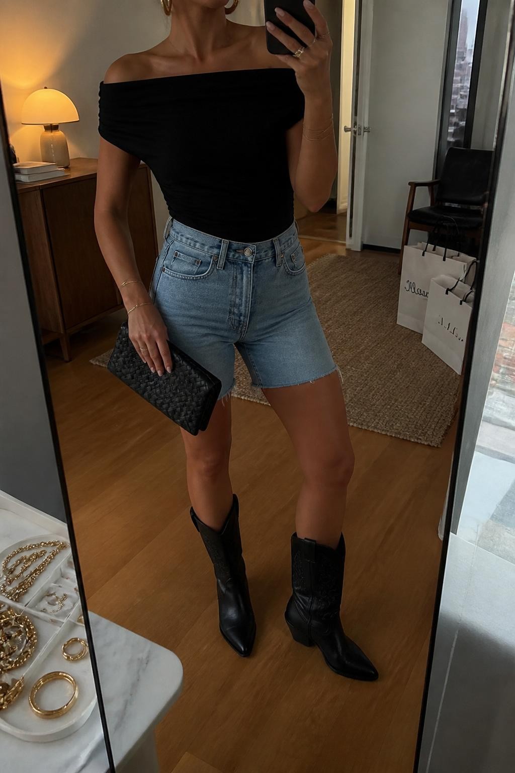 mirror selfie of a woman wearing light blue high-rise denim jorts, a black off-shoulder top, black leather cowboy boots, black woven clutch, and chunky gold hoop earrings, clearly showing the same clothing items, colors, fabrics, and accessories listed in the Outfit Pieces section, holding a phone in front of a mirror, cropped at NECK so the face does not show, with a relaxed casual selfie pose, in a stylish city apartment, warm moody lighting, including a jewelry tray on the vanity and shopping bags beside a chair, latest iPhone photo quality.