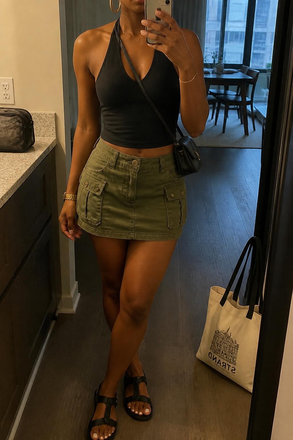 Outfit Image Prompt:
mirror selfie of a Black woman with warm brown skin wearing a black fitted halter top, olive green cargo mini skirt, black sporty sandals, small black crossbody bag, and chunky gold hoop earrings, holding a phone in front of a mirror, cropped at NECK so the face does not show, relaxed NYC summer street style selfie pose, in a stylish city apartment, warm moody lighting, with a tote bag resting on the floor and a makeup bag on the counter, realistic latest iPhone photo quality, natural skin texture.