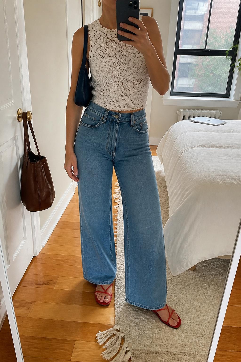 Image Prompt:
mirror selfie of a woman wearing a white crochet sleeveless top with a lined front, high-waisted medium-wash wide-leg jeans, red flat sandals, a small navy shoulder bag, and gold drop earrings, clearly showing the same clothing items, colors, fabrics, and accessories listed in the Outfit Pieces section, holding a phone in front of a mirror, cropped at NECK so the face does not show, with a relaxed casual selfie pose, in a stylish city apartment, natural window light, with a handbag hanging on a door handle and a laptop or tablet on the bed, natural casual mirror selfie, not staged, latest iPhone photo quality.