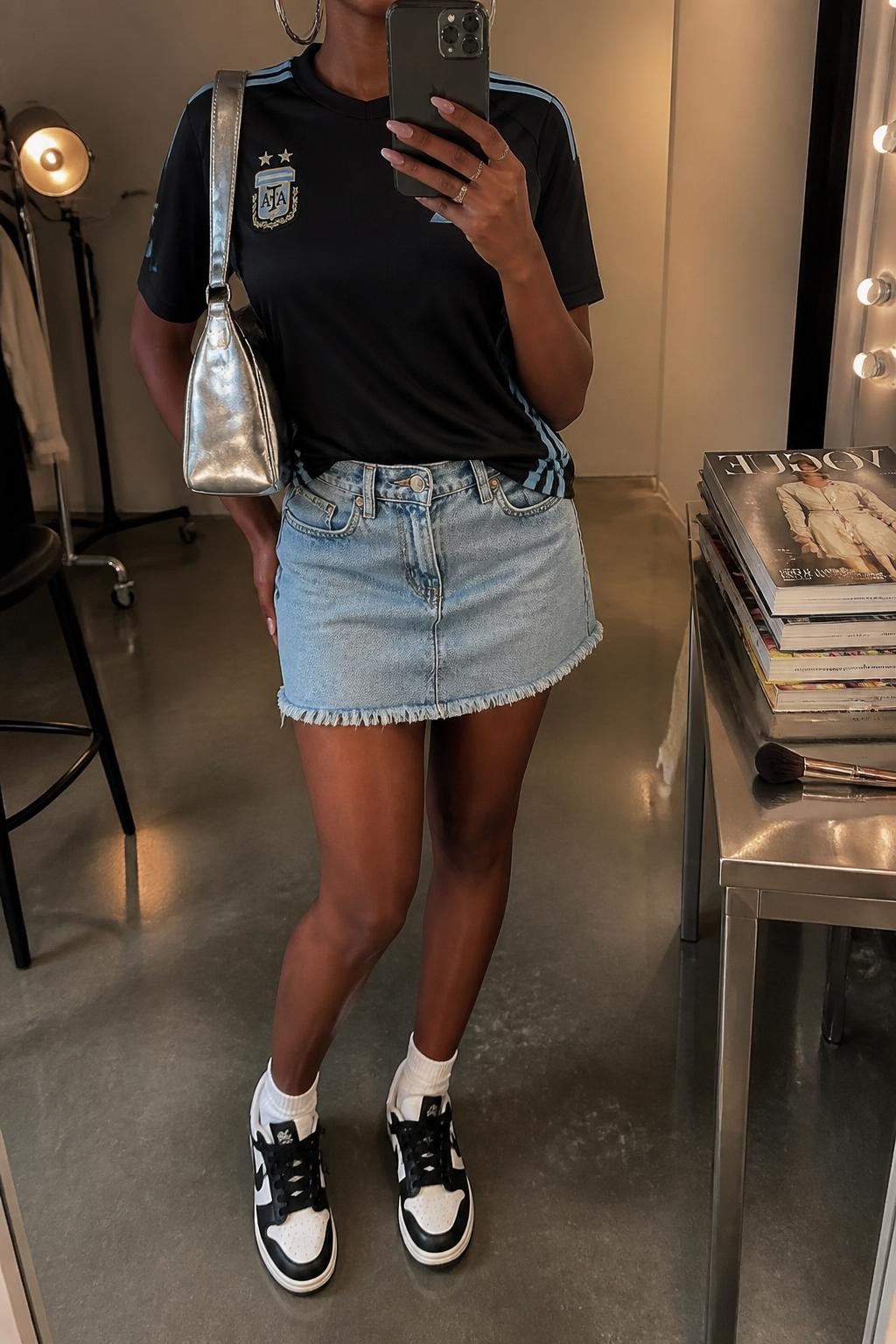 mirror selfie of a Black woman wearing a black Argentina soccer jersey, light blue denim mini skirt, black and white sneakers, silver shoulder bag, and chunky silver hoop earrings, clearly showing the same clothing items, colors, fabrics, and accessories listed in the Outfit Pieces section, holding a phone in front of a mirror, cropped at NECK so the face does not show, with a relaxed casual selfie pose, in a fashion studio mirror corner, warm moody lighting, including a stack of fashion magazines on a side table and a makeup brush on the counter, latest iPhone photo quality.