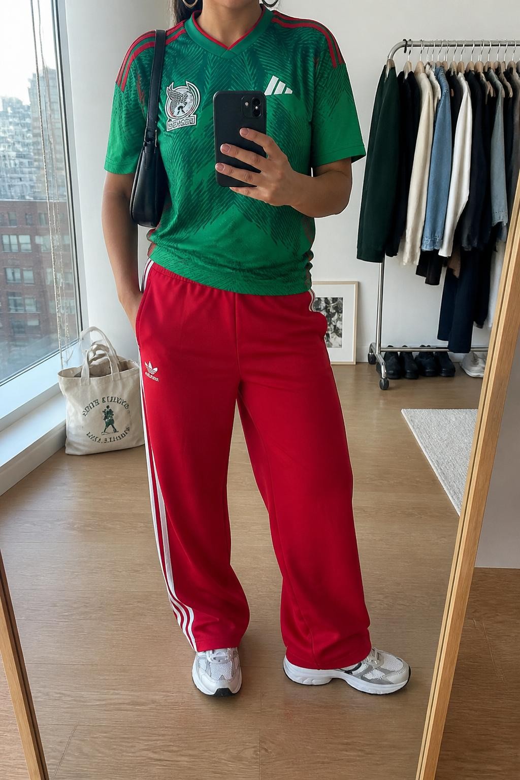 mirror selfie of a woman wearing a green Mexico soccer jersey, red relaxed track pants with white side stripes, white running sneakers, black mini shoulder bag, and silver hoop earrings, clearly showing the same clothing items, colors, fabrics, and accessories listed in the Outfit Pieces section, holding a phone in front of a mirror, cropped at NECK so the face does not show, with a relaxed casual selfie pose, in a stylish city apartment, natural window light, including a tote bag resting on the floor and a clothing rack with garments behind, latest iPhone photo quality.