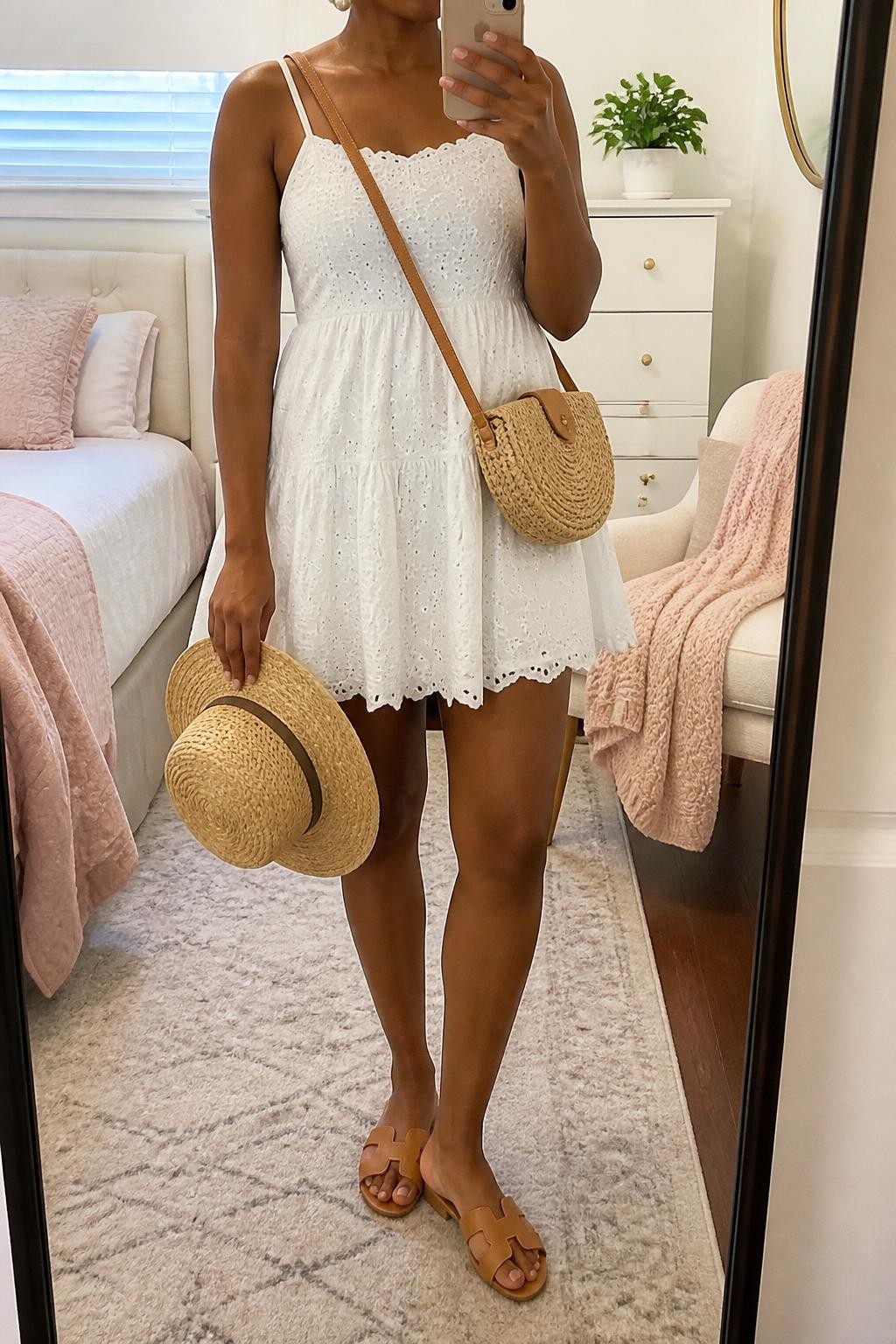 mirror selfie of a Black woman wearing a white eyelet mini dress, tan flat slide sandals, woven straw sun hat held in one hand, tan raffia crossbody bag, and pearl stud earrings, clearly showing the same clothing items, colors, fabrics, and accessories listed in the Outfit Pieces section, holding a phone in front of a mirror, cropped at NECK so the face does not show, with a relaxed casual selfie pose, in a feminine-themed bedroom, natural window light, including a folded blanket on a chair and a small plant on a dresser, latest iPhone photo quality.