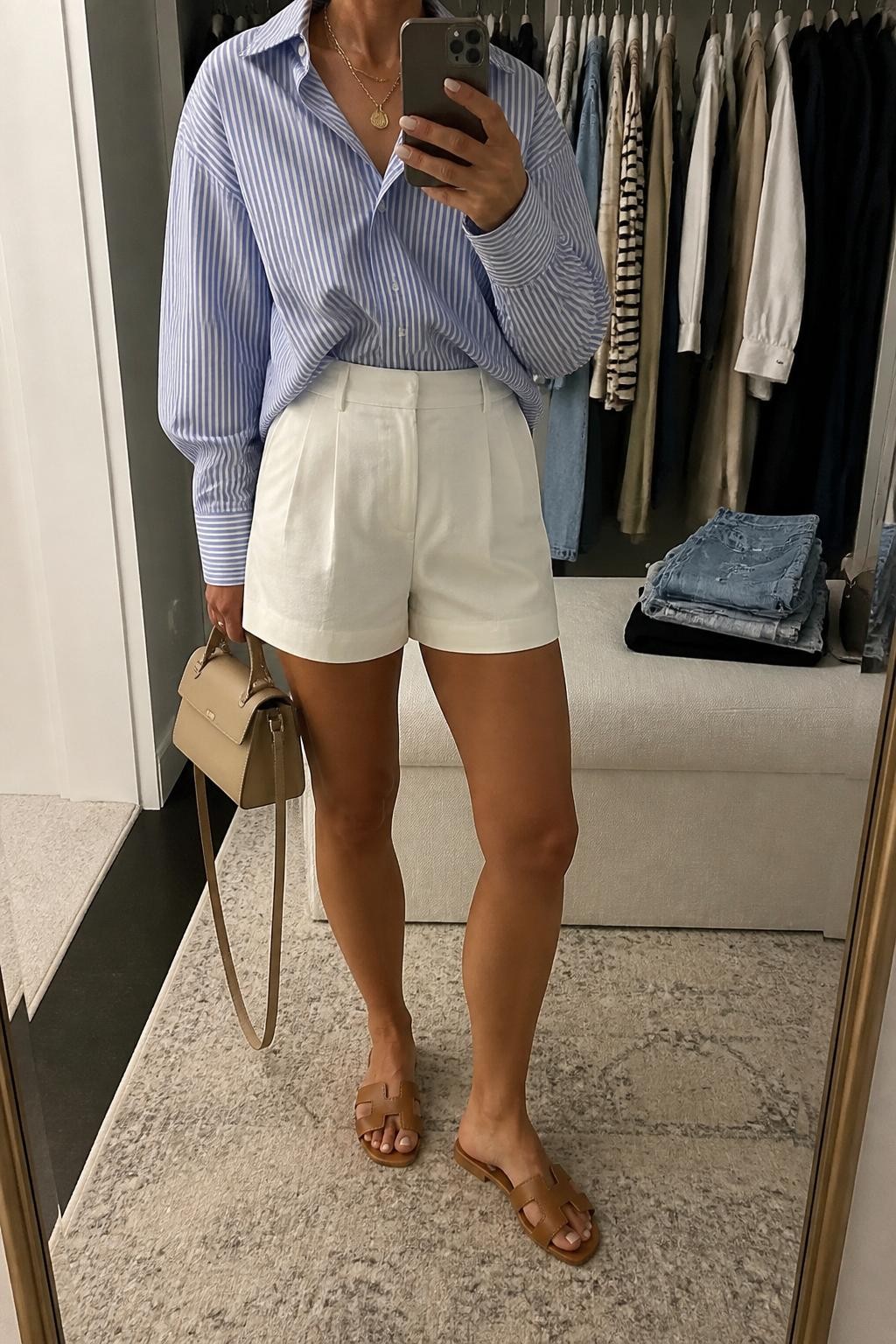 Outfit Image Prompt:
mirror selfie of a woman wearing a blue and white striped oversized cotton button-down shirt, white high-waisted tailored shorts, tan leather slide sandals, a structured beige mini bag, and gold layered necklaces, holding a phone in front of a mirror, cropped at NECK so the face does not show, relaxed casual selfie pose, in a walk-in closet mirror, soft indoor lighting, with a clothing rack with garments behind and folded denim jeans on a bench, realistic latest iPhone photo quality, natural casual mirror selfie.