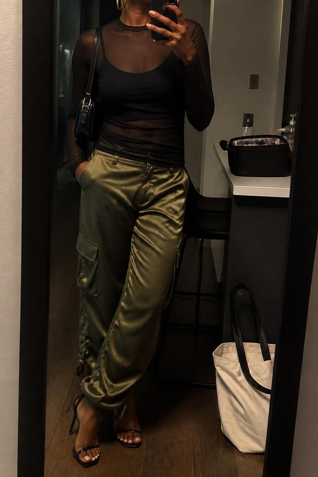 Outfit Image Prompt:
mirror selfie of a Black woman with warm brown skin wearing a sheer black long-sleeve layered top over a black fitted camisole, olive satin cargo pants, black strappy heeled sandals, black mini shoulder bag, and chunky gold hoop earrings, holding a phone in front of a mirror, cropped at NECK so the face does not show, relaxed high-fashion city selfie pose, in a stylish city apartment, warm moody lighting, with a makeup bag on the counter and a tote bag resting on the floor, natural skin texture, realistic latest iPhone photo quality.