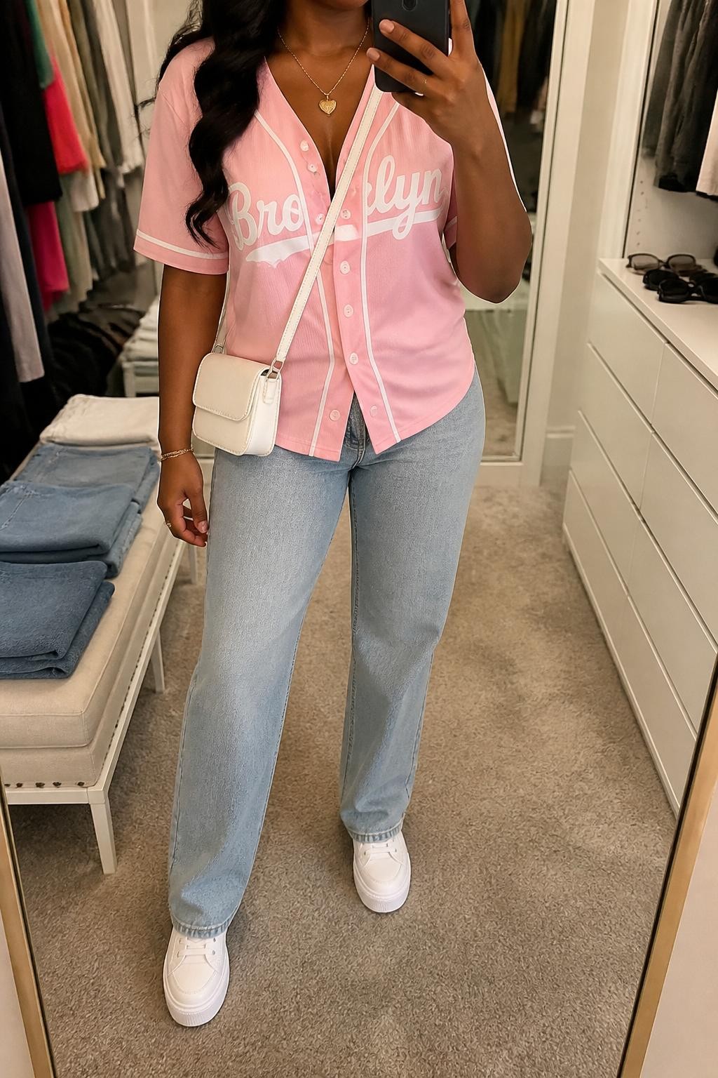 mirror selfie of a Black woman wearing a pink button-front baseball jersey, light blue straight-leg jeans, white platform sneakers, white mini crossbody bag, and gold heart pendant necklace, clearly showing the same clothing items, colors, fabrics, and accessories listed in the Outfit Pieces section, holding a phone in front of a mirror, cropped at NECK so the face does not show, with a relaxed casual selfie pose, in a walk-in closet mirror, soft indoor lighting, including folded denim jeans on a bench and sunglasses placed on a dresser, latest iPhone photo quality.