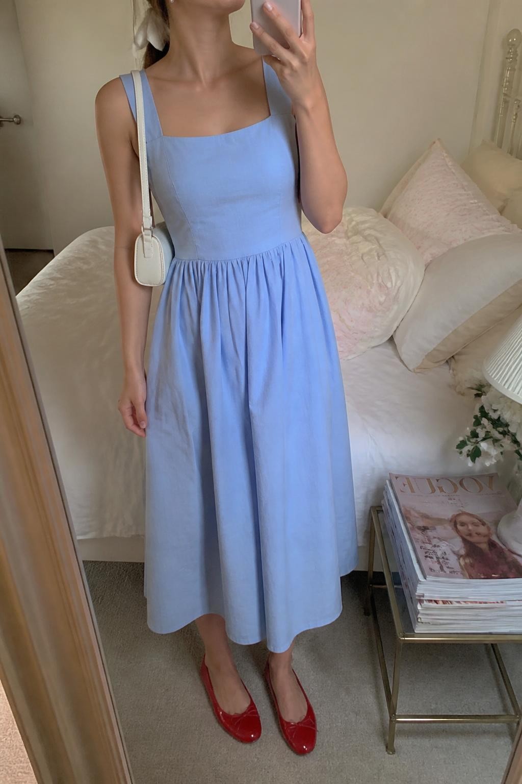 Outfit Image Prompt:
mirror selfie of a woman wearing a blue cotton midi dress with a square neckline, red ballet flats, a small white shoulder bag, pearl stud earrings, and a white ribbon tied around a low ponytail cropped at neck, holding a phone in front of a mirror, cropped at NECK so the face does not show, relaxed casual selfie pose, in a feminine-themed bedroom, soft indoor lighting, with soft pillows on an unmade bed and a stack of fashion magazines on a side table, realistic latest iPhone photo quality, soft romantic styling.