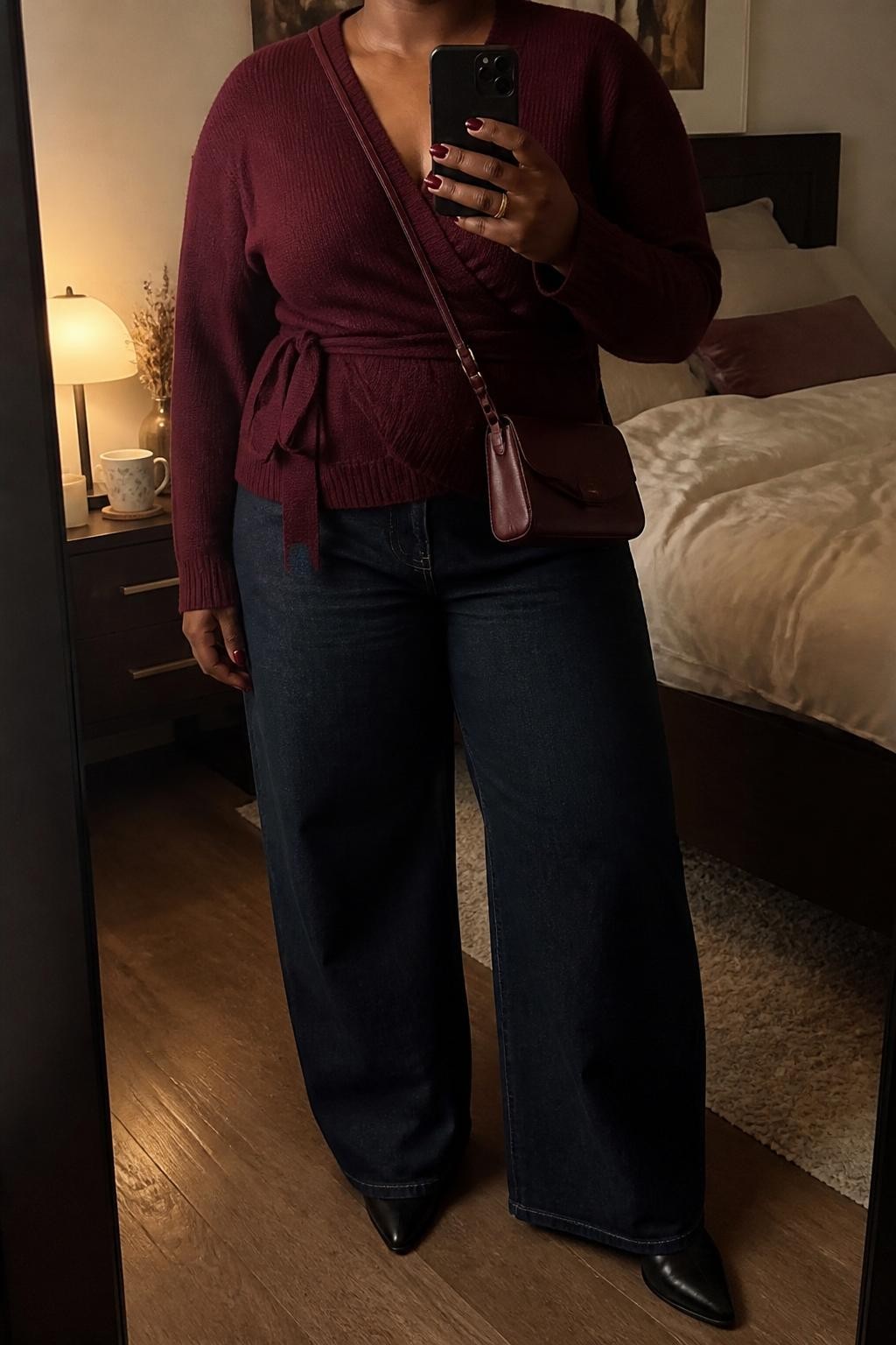 mirror selfie of a plus size Black woman wearing the exact outfit described above, a deep burgundy wrap sweater with a soft knit texture, dark indigo wide-leg jeans, black pointed ankle boots, burgundy faux leather mini crossbody bag, and soft berry lip color visible below the neck crop only, clearly showing the same clothing items, colors, fabrics, and accessories listed in the Outfit Pieces section, holding a phone in front of a mirror, cropped at NECK so the face does not show, with a relaxed casual selfie pose, in a cozy bedroom mirror setup, warm moody lighting, including a coffee mug on a bedside table and soft pillows on an unmade bed, latest iPhone photo quality.