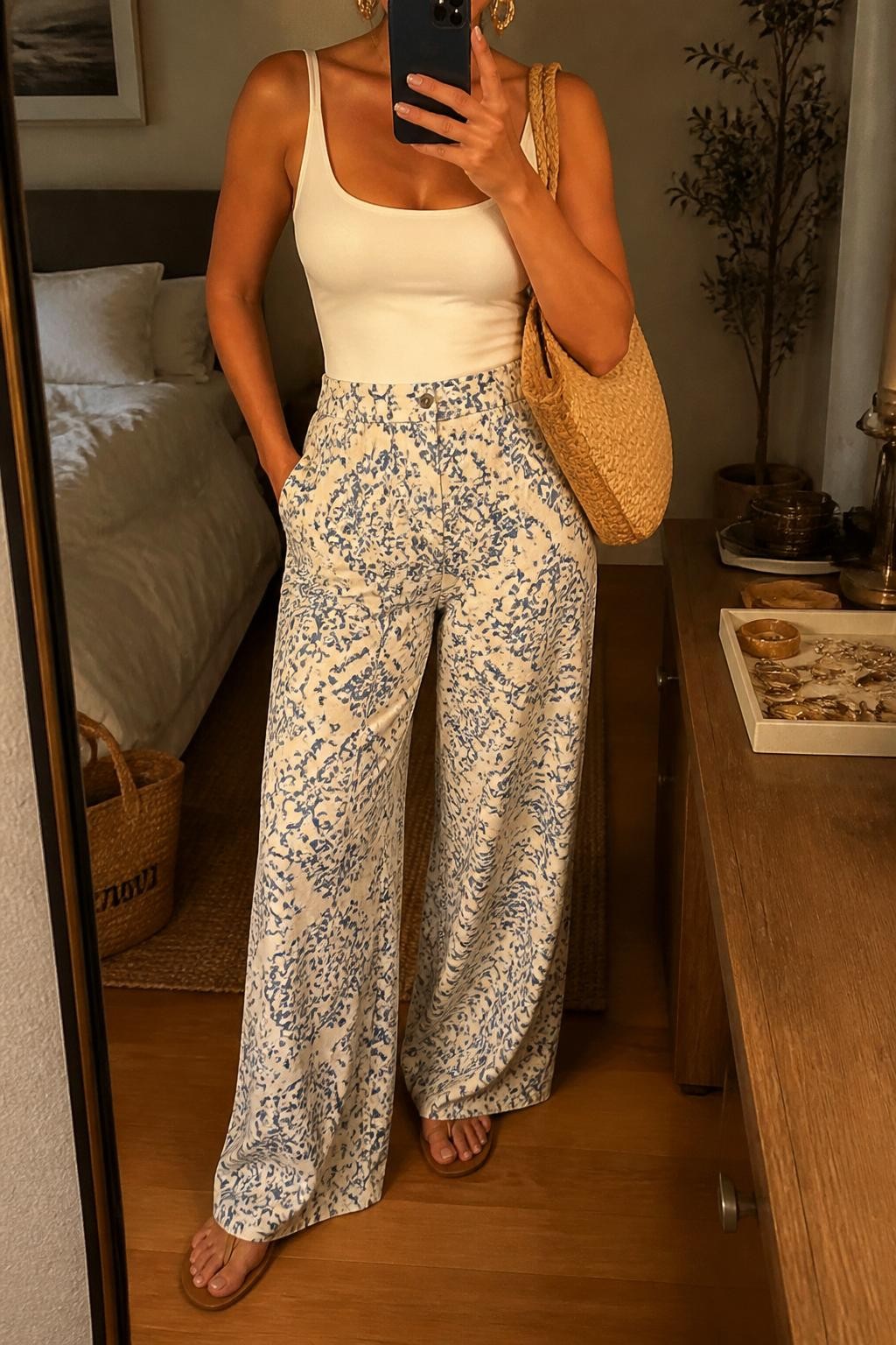 Outfit Image Prompt:
mirror selfie of a woman wearing a fitted white tank bodysuit, blue and cream printed palazzo pants, tan flat sandals, a beige raffia tote bag, and chunky gold earrings, holding a phone in front of a mirror, cropped at NECK so the face does not show, relaxed casual selfie pose, in a cozy bedroom mirror setup, evening golden hour lighting, with a tote bag resting on the floor and a jewelry tray on the vanity, realistic latest iPhone photo quality, relaxed summer travel styling.