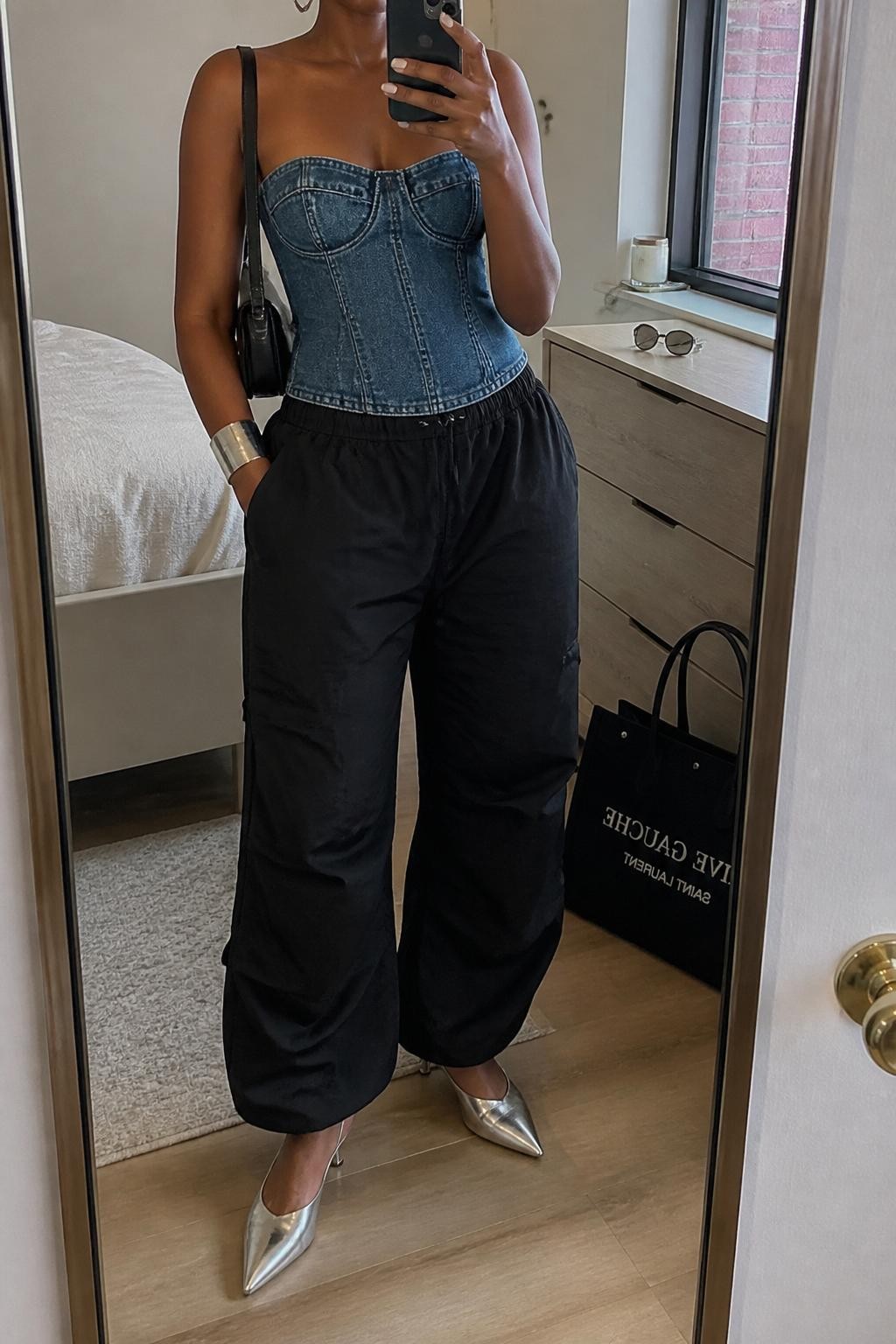 Outfit Image Prompt:
mirror selfie of a Black woman with medium brown skin wearing a structured denim bustier top, black relaxed parachute pants, silver pointed kitten heels, small black shoulder bag, chunky silver cuff bracelet, and silver hoop earrings, holding a phone in front of a mirror, cropped at NECK so the face does not show, relaxed downtown fashion selfie pose, in a cozy bedroom mirror setup, natural window light, with a tote bag resting on the floor and sunglasses placed on a dresser, natural skin texture, realistic latest iPhone photo quality.