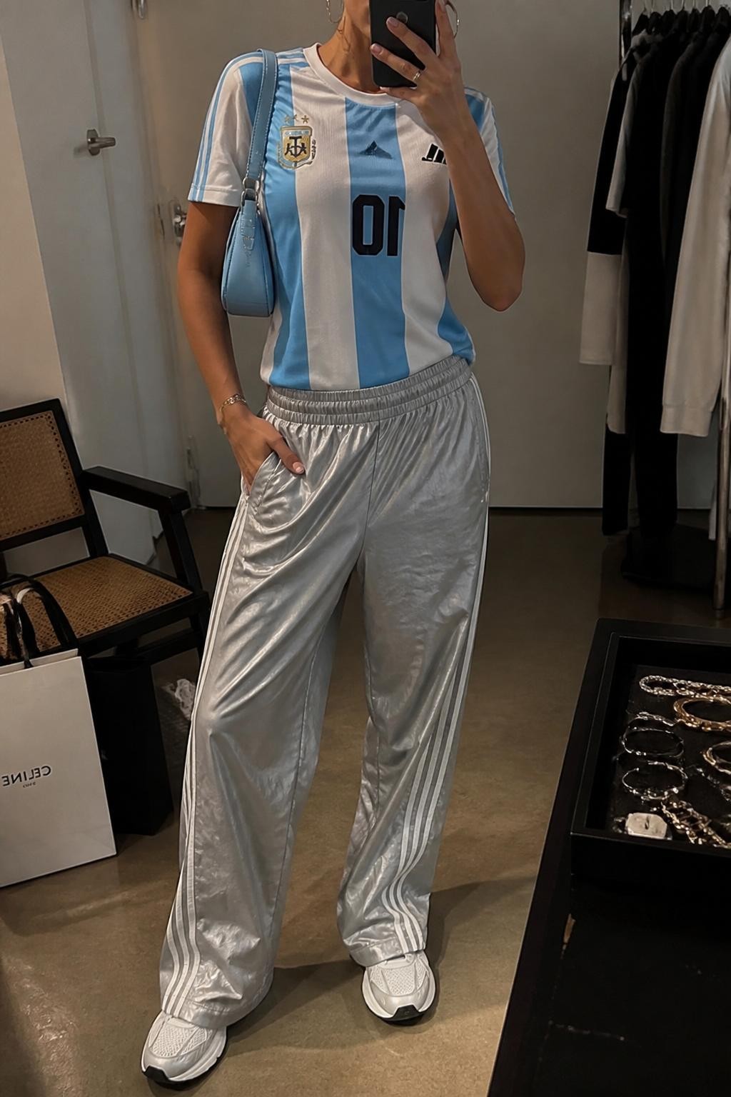 mirror selfie of a woman wearing a sky blue and white Argentina soccer jersey, silver relaxed track pants with white side stripes, white running sneakers, pale blue mini shoulder bag, and silver hoop earrings, clearly showing the same clothing items, colors, fabrics, and accessories listed in the Outfit Pieces section, holding a phone in front of a mirror, cropped at NECK so the face does not show, with a relaxed casual selfie pose, in a fashion studio mirror corner, warm moody lighting, including a jewelry tray on the vanity and shopping bags beside a chair, latest iPhone photo quality.