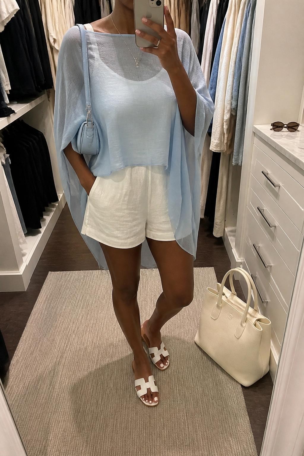 mirror selfie of a Black woman wearing a sky blue sheer poncho layered over a white sleeveless linen romper, white flat sandals, pale blue mini shoulder bag, and delicate silver necklace, clearly showing the same clothing items, colors, fabrics, and accessories listed in the Outfit Pieces section, holding a phone in front of a mirror, cropped at NECK so the face does not show, with a relaxed casual selfie pose, in a walk-in closet mirror, soft indoor lighting, including sunglasses placed on a dresser and a cream tote bag resting on the floor, latest iPhone photo quality.