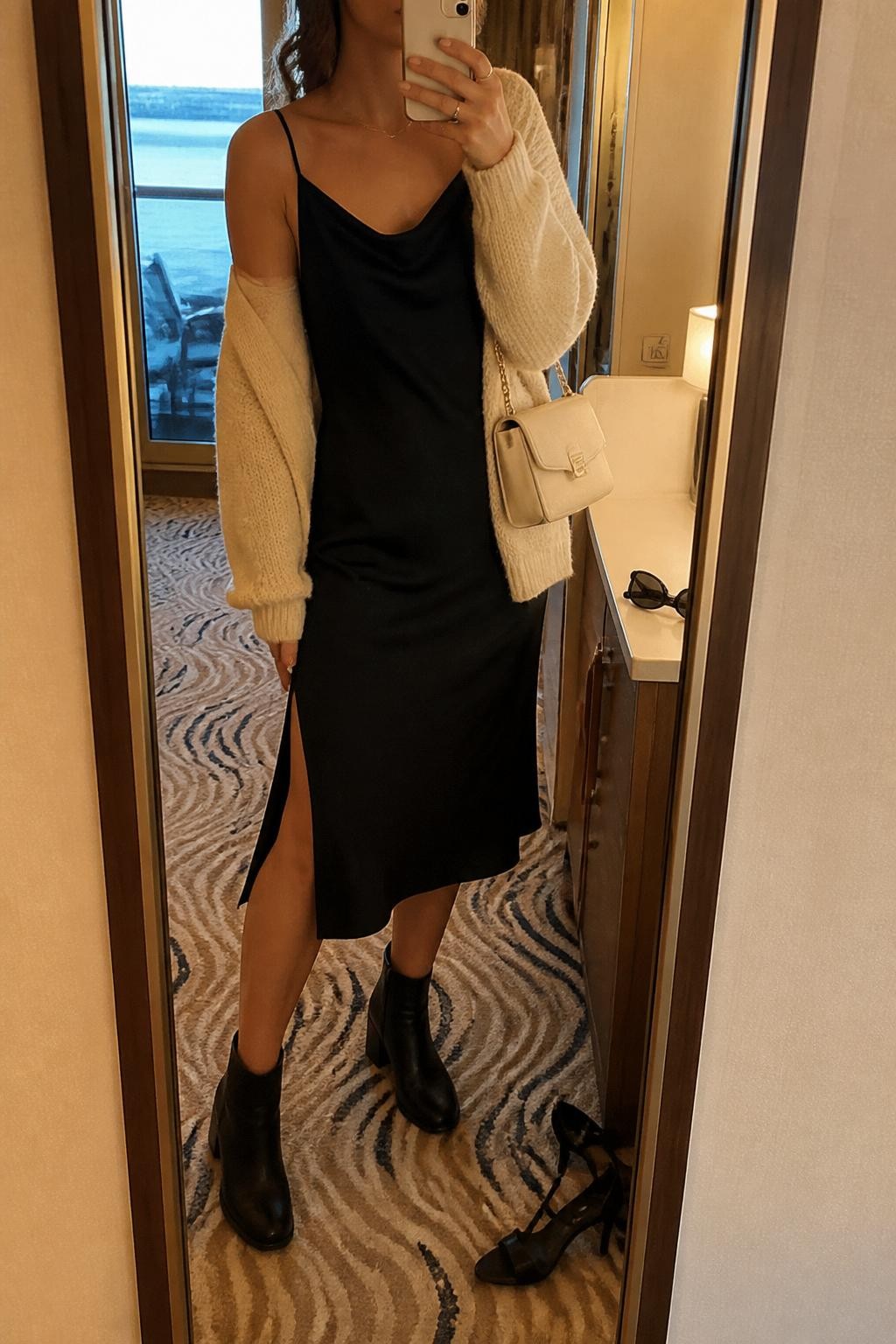 Outfit Image Prompt:
mirror selfie of a woman wearing a black satin slip dress, cream chunky knit cardigan, black block-heel ankle boots, a pearl hair clip, and a small cream shoulder bag, holding a phone in front of a mirror, cropped at NECK so the face does not show, relaxed casual selfie pose, in a stylish cruise cabin mirror corner with warm moody lighting, including a pair of heels on the floor nearby and sunglasses placed on a dresser, natural skin texture, realistic latest iPhone photo quality, soft dinner outfit styling.
