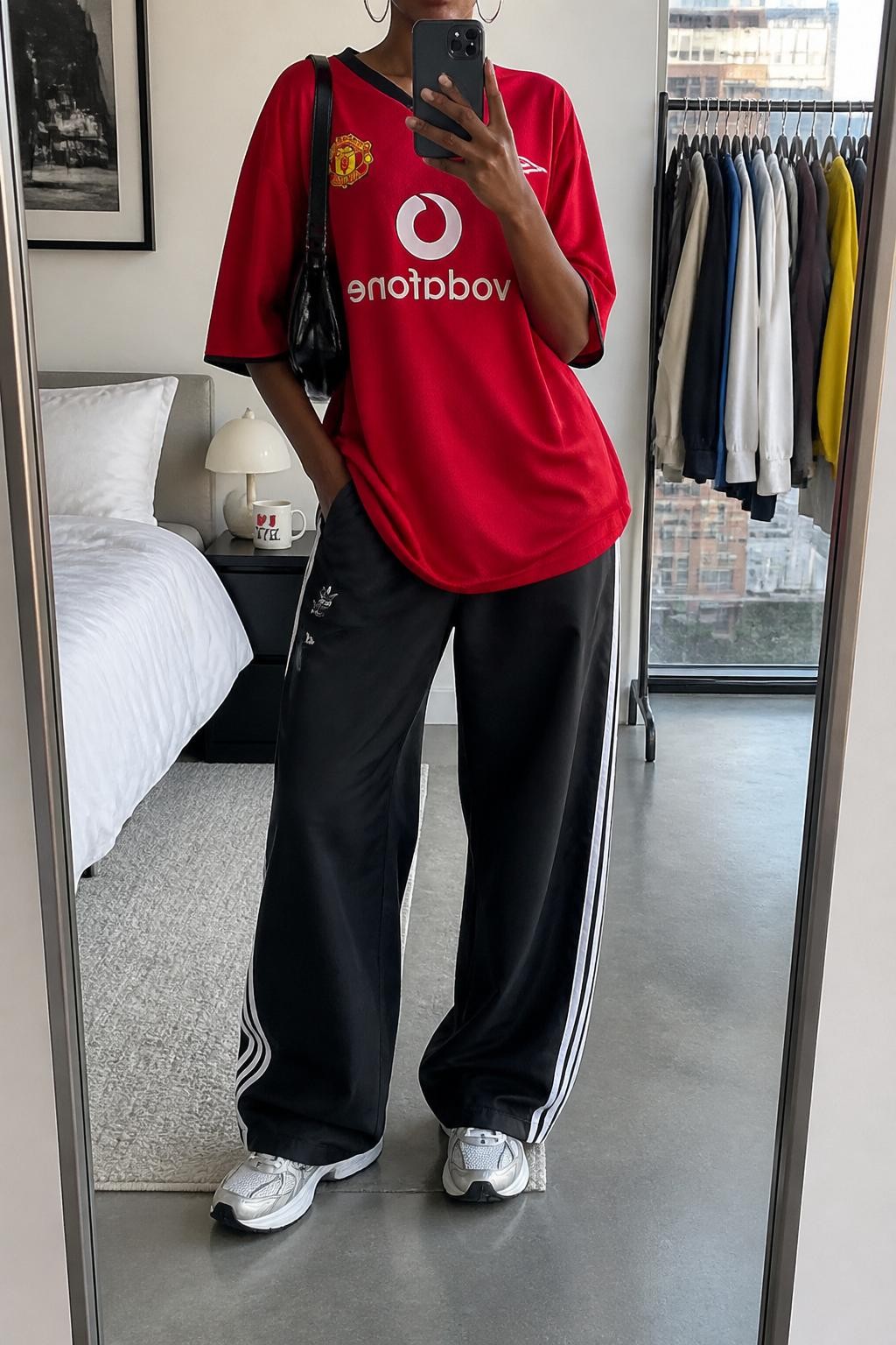 mirror selfie of a Black woman wearing an oversized red soccer jersey, black relaxed track pants with white side stripes, white running sneakers, black mini shoulder bag, and silver hoop earrings, clearly showing the same clothing items, colors, fabrics, and accessories listed in the Outfit Pieces section, holding a phone in front of a mirror, cropped at NECK so the face does not show, with a relaxed casual selfie pose, in a stylish city apartment, natural window light, including a clothing rack with garments behind and a coffee mug on a bedside table, latest iPhone photo quality.