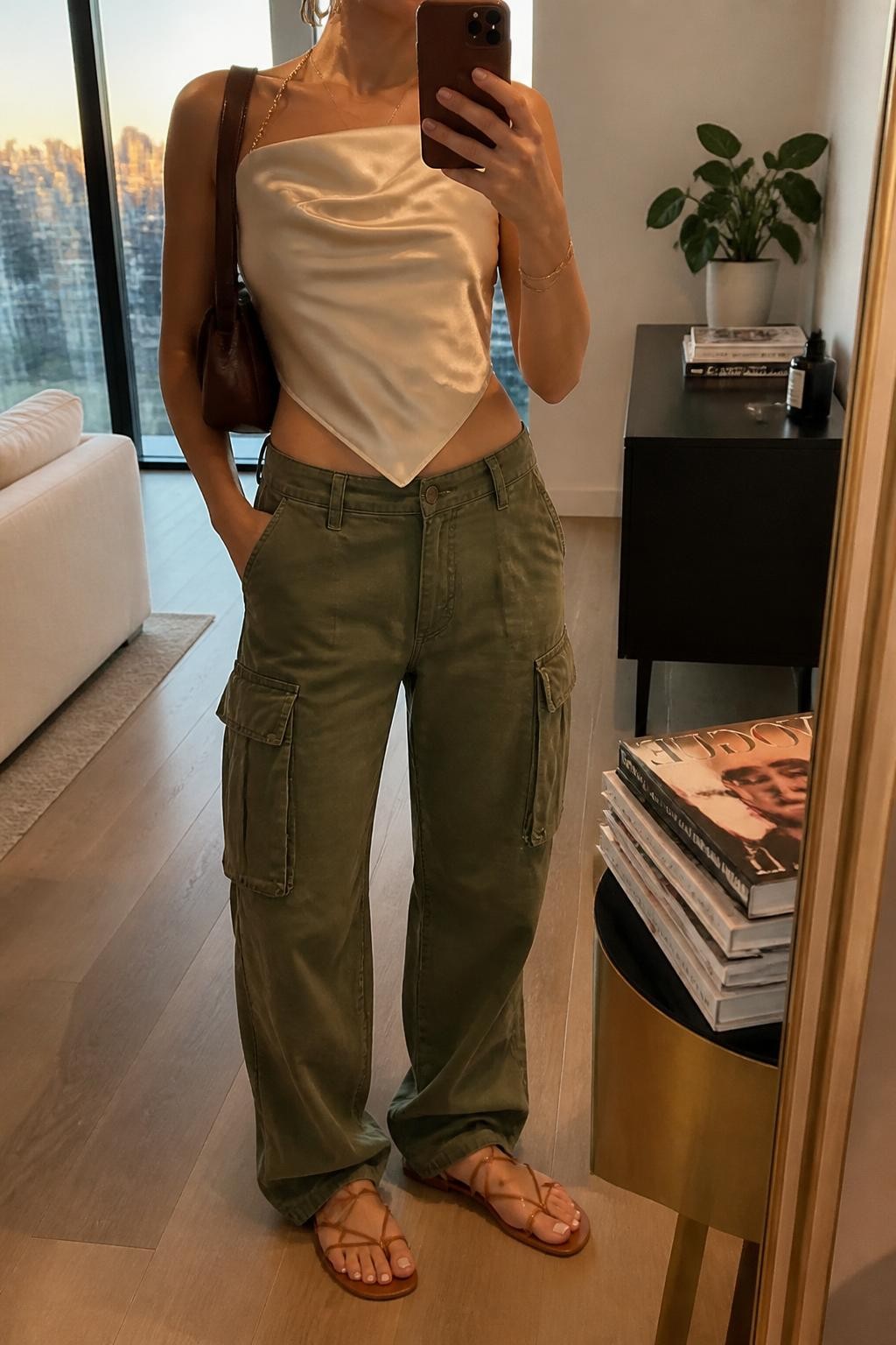mirror selfie of a woman wearing olive green cargo pants, cream silk scarf top, tan lace-up flat sandals, brown mini shoulder bag, and gold hoop earrings, clearly showing the same clothing items, colors, fabrics, and accessories listed in the Outfit Pieces section, holding a phone in front of a mirror, cropped at NECK so the face does not show, with a relaxed casual selfie pose, in a stylish city apartment, evening golden hour, including a stack of fashion magazines on a side table and a small plant on a dresser, latest iPhone photo quality.