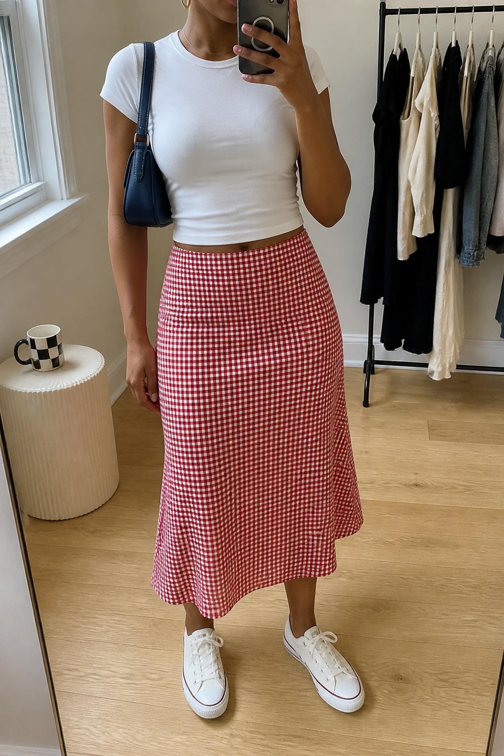 Outfit Image Prompt:
mirror selfie of a Black woman with medium brown skin wearing a fitted white baby tee, red gingham midi skirt, white canvas sneakers, a small navy shoulder bag, and gold huggie earrings, clearly showing the exact clothing items and accessories listed, holding a phone in front of a mirror, cropped at NECK so the face does not show, relaxed casual selfie pose, in a minimal dressing room, natural window light, with a clothing rack with garments behind and a coffee mug on a side table, natural skin texture, latest iPhone photo quality, realistic mirror selfie.