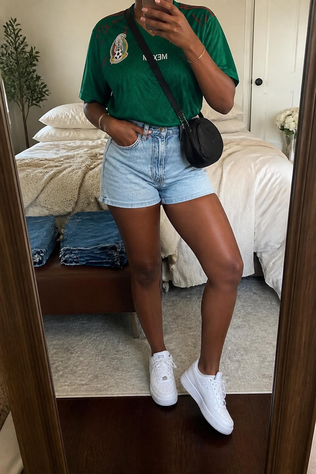 mirror selfie of a Black woman wearing an oversized green Mexico soccer jersey, light wash high-rise denim shorts, white low-top sneakers, black mini crossbody bag, and red baseball cap, clearly showing the same clothing items, colors, fabrics, and accessories listed in the Outfit Pieces section, holding a phone in front of a mirror, cropped at NECK so the face does not show, with a relaxed casual selfie pose, in a cozy bedroom mirror setup, soft indoor lighting, including soft pillows on an unmade bed and folded denim jeans on a bench, latest iPhone photo quality.