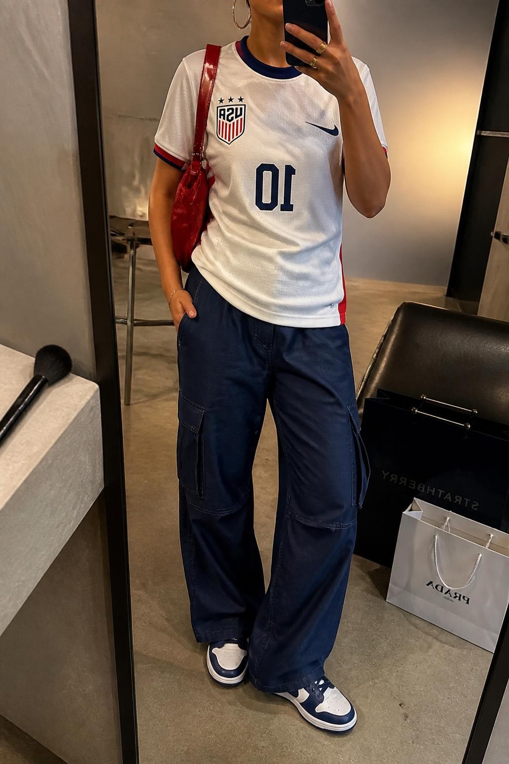 mirror selfie of a woman wearing a white USA soccer jersey, navy relaxed cargo pants, white and navy retro sneakers, red nylon shoulder bag, and chunky silver hoop earrings, clearly showing the same clothing items, colors, fabrics, and accessories listed in the Outfit Pieces section, holding a phone in front of a mirror, cropped at NECK so the face does not show, with a relaxed casual selfie pose, in a fashion studio mirror corner, warm moody lighting, including a makeup brush on the counter and shopping bags beside a chair, latest iPhone photo quality.