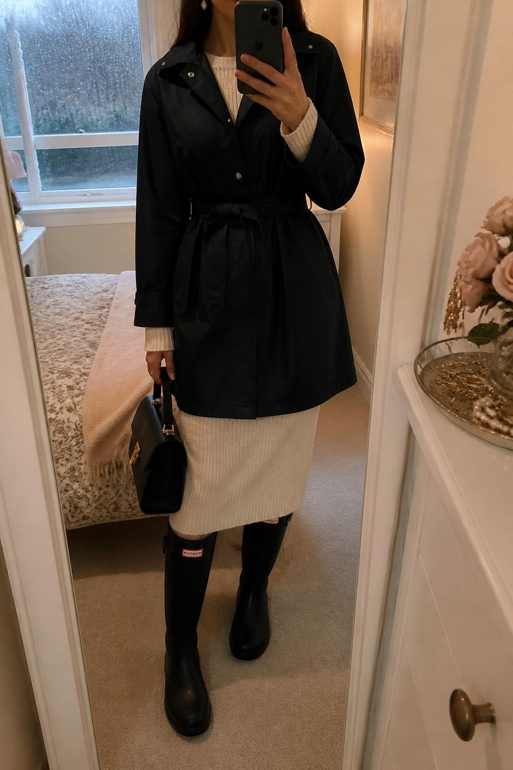 mirror selfie of a woman wearing the exact outfit described above, clearly showing a black belted raincoat layered over a cream knit sweater and matching cream knit midi skirt, black knee-high rain boots, a black top-handle bag, and pearl earrings, holding a phone in front of a mirror, cropped at NECK so the face does not show, with a relaxed casual selfie pose, in a feminine bedroom mirror setup, soft indoor lighting, with rain visible through the window and a jewelry tray on the vanity, latest iPhone photo quality.