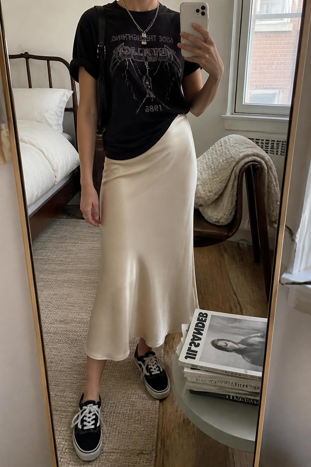 Outfit Image Prompt:
mirror selfie of a woman wearing a vintage-style black graphic tee, champagne satin midi skirt, black low-top sneakers, black leather shoulder bag, and silver layered necklaces, holding a phone in front of a mirror, cropped at NECK so the face does not show, relaxed casual city selfie pose, in a cozy bedroom mirror setup, natural window light, with folded blanket on a chair and a stack of fashion magazines on a side table, realistic latest iPhone photo quality, effortless NYC street style.