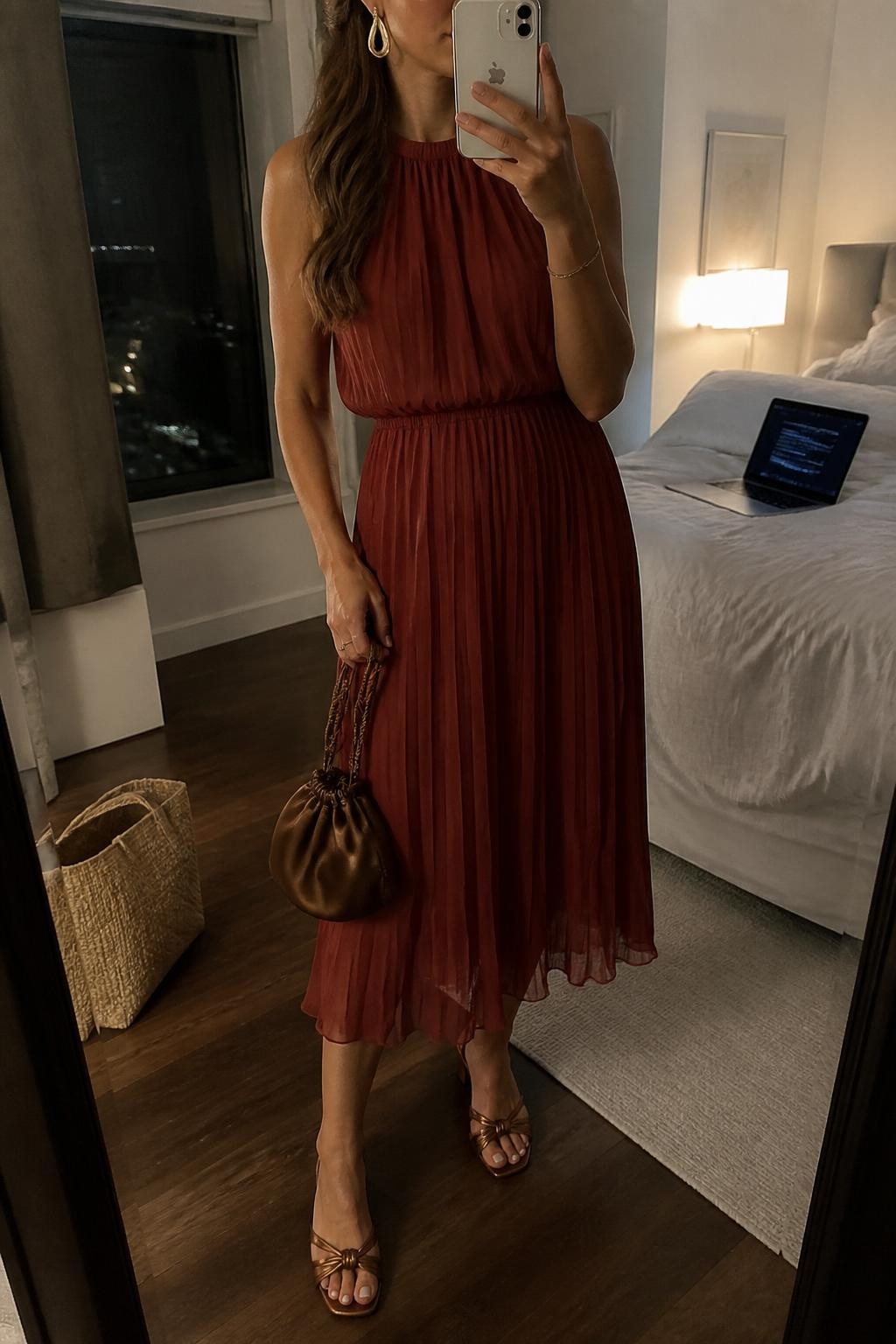 mirror selfie of a woman wearing a brick red pleated chiffon midi cocktail dress, bronze heeled sandals, gold teardrop earrings, a bronze satin pouch bag, and half-up waves with peachy makeup, clearly showing the same clothing items, colors, fabrics, and accessories listed in the Outfit Pieces section, holding a phone in front of a mirror, cropped at NECK so the face does not show, with a relaxed casual selfie pose, in a stylish city apartment, warm moody lighting, including laptop or tablet on the bed and tote bag resting on the floor, latest iPhone photo quality.