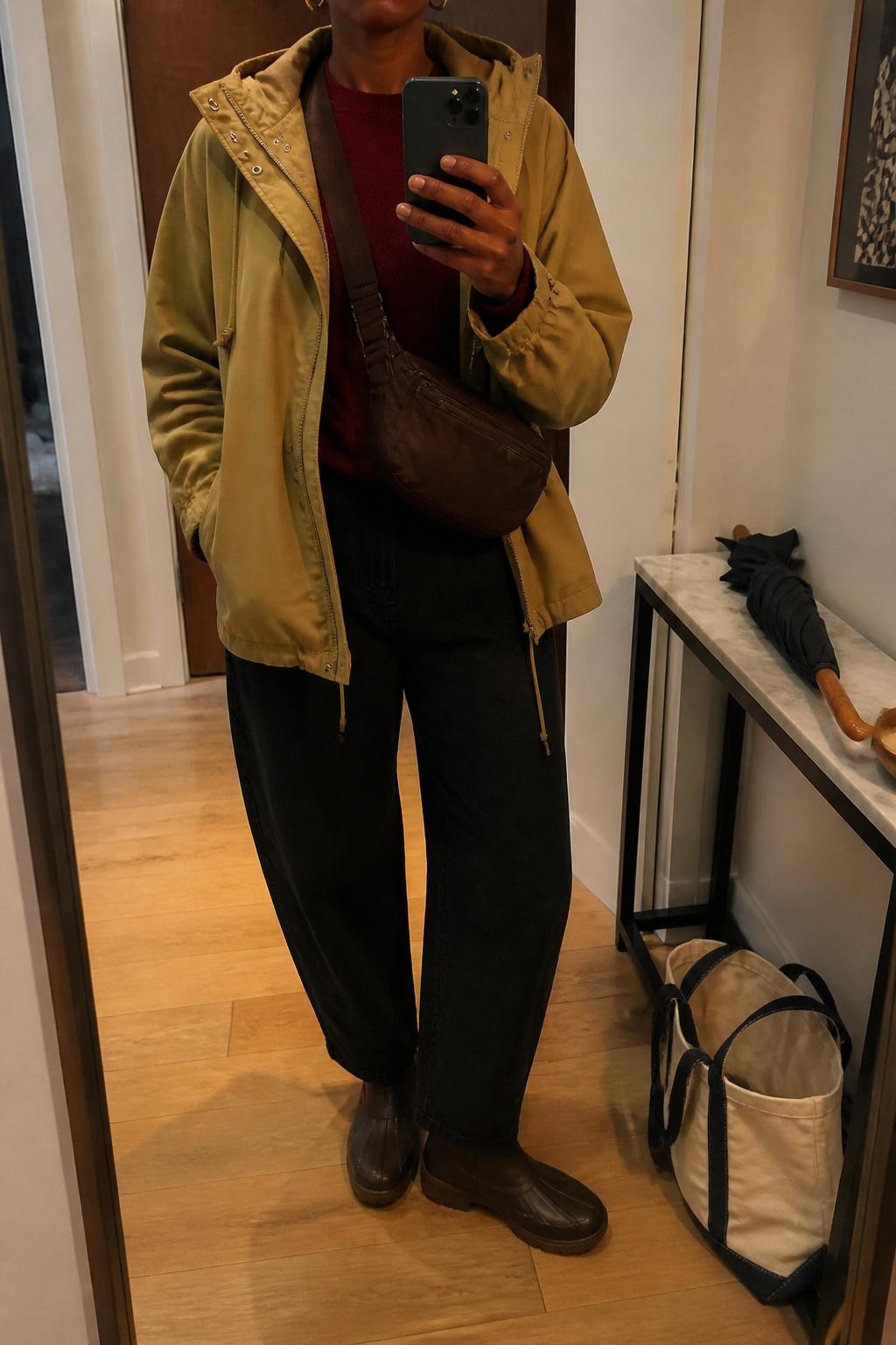 mirror selfie of a Black woman wearing the exact outfit described above, clearly showing a tan hooded anorak layered over a burgundy crewneck sweater, black barrel jeans, dark brown waterproof ankle boots, a brown nylon crossbody bag, and small gold hoop earrings, holding a phone in front of a mirror, cropped at NECK so the face does not show, with a relaxed casual selfie pose, in a modern apartment hallway mirror, warm indoor lighting, with a folded umbrella on a console table and a tote bag on the floor, latest iPhone photo quality.