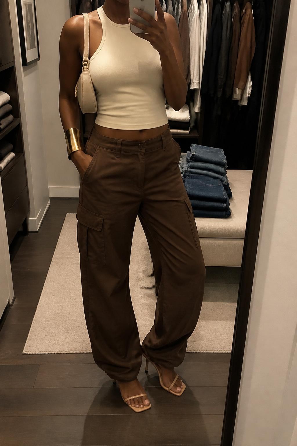 mirror selfie of a Black woman wearing chocolate brown cargo pants, cream fitted halter top, tan strappy heeled sandals, cream mini shoulder bag, and gold cuff bracelet, clearly showing the same clothing items, colors, fabrics, and accessories listed in the Outfit Pieces section, holding a phone in front of a mirror, cropped at NECK so the face does not show, with a relaxed casual selfie pose, in a walk-in closet mirror, warm moody lighting, including an open closet with clothes visible and folded denim jeans on a bench, latest iPhone photo quality.