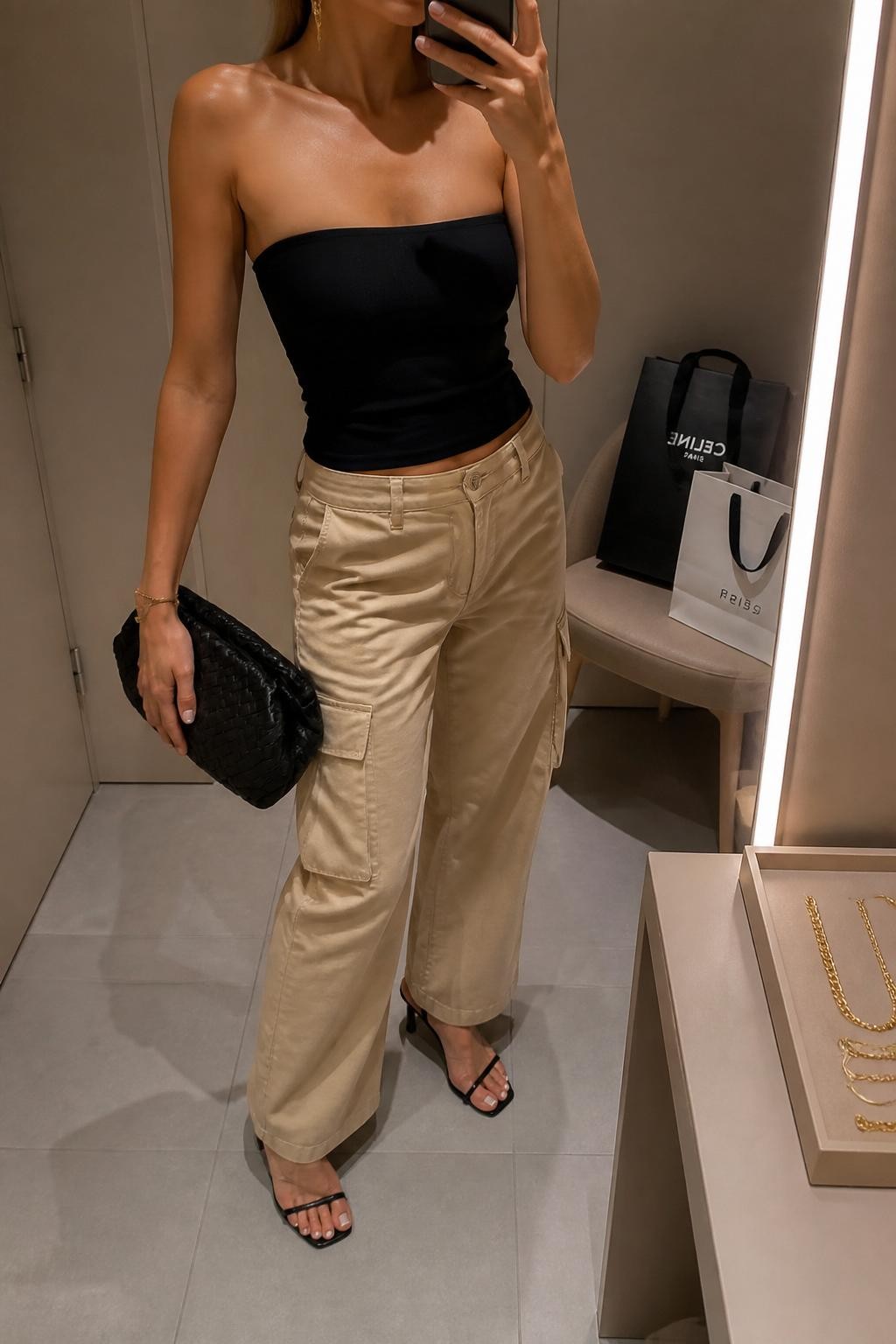 mirror selfie of a woman wearing sand-colored cargo pants, fitted black strapless top, black kitten-heel sandals, black woven clutch, and gold drop earrings, clearly showing the same clothing items, colors, fabrics, and accessories listed in the Outfit Pieces section, holding a phone in front of a mirror, cropped at NECK so the face does not show, with a relaxed casual selfie pose, in a minimal dressing room, evening golden hour, including a jewelry tray on the vanity and shopping bags beside a chair, latest iPhone photo quality.