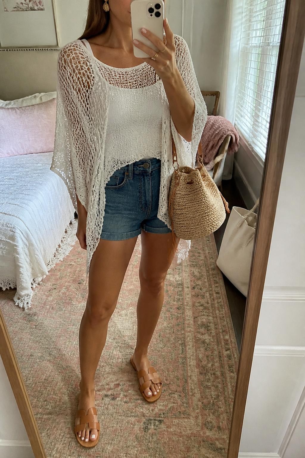 mirror selfie of a woman wearing a white crochet sheer poncho layered over a white fitted tank top, medium blue high-rise denim shorts, tan leather flat sandals, raffia bucket bag, and gold shell earrings, clearly showing the same clothing items, colors, fabrics, and accessories listed in the Outfit Pieces section, holding a phone in front of a mirror, cropped at NECK so the face does not show, with a relaxed casual selfie pose, in a feminine-themed bedroom, natural window light, including a cream tote bag resting on the floor and a folded blanket on a chair, latest iPhone photo quality.