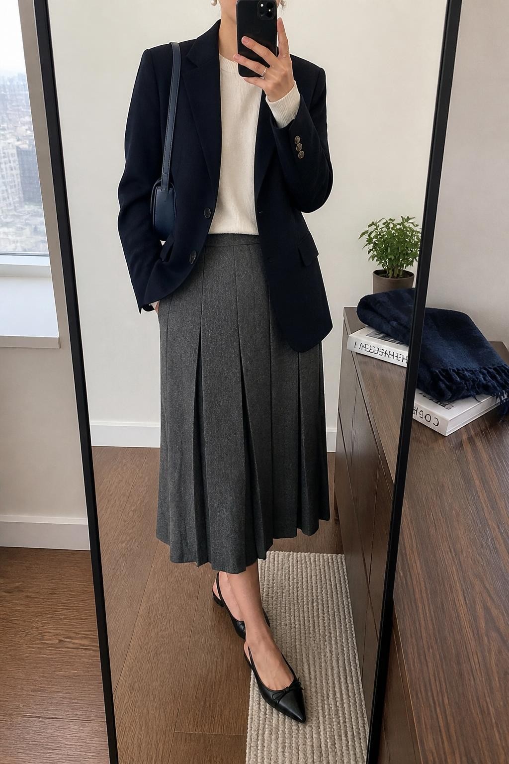 mirror selfie of a woman wearing the exact outfit described above, clearly showing a navy wool blazer layered over an ivory fine-knit sweater, gray pleated midi skirt, black slingback flats, a navy leather shoulder bag, and pearl stud earrings, holding a phone in front of a mirror, cropped at NECK so the face does not show, with a relaxed casual selfie pose, in a modern apartment-style mirror, natural window light, with a small plant on the dresser and folded scarf beside a travel guidebook, latest iPhone photo quality.