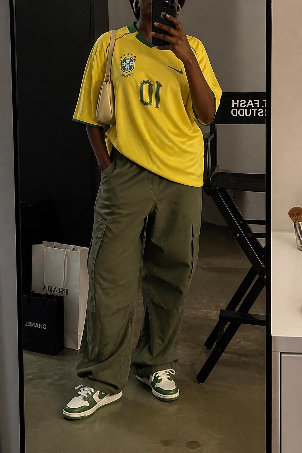 mirror selfie of a Black woman wearing an oversized yellow soccer jersey, olive green relaxed cargo pants, white and green retro sneakers, beige nylon shoulder bag, and small gold hoop earrings, clearly showing the same clothing items, colors, fabrics, and accessories listed in the Outfit Pieces section, holding a phone in front of a mirror, cropped at NECK so the face does not show, with a relaxed casual selfie pose, in a fashion studio mirror corner, warm moody lighting, including a makeup brush on the counter and shopping bags beside a chair, latest iPhone photo quality.
