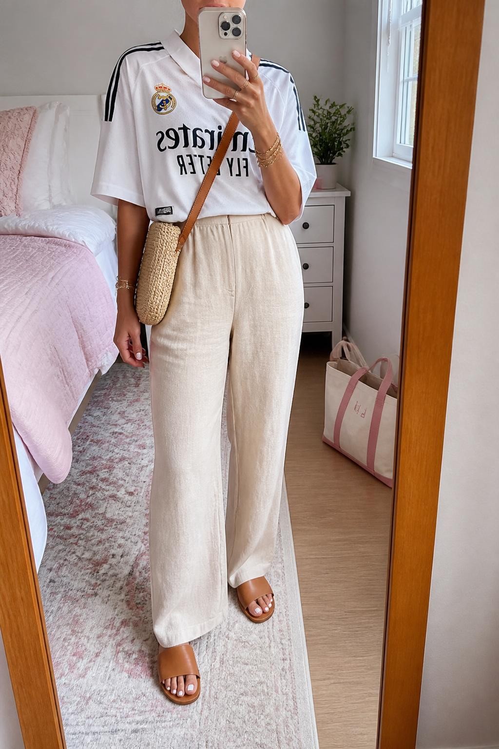 mirror selfie of a woman wearing an oversized white soccer jersey, cream high-waisted linen pants, tan leather slide sandals, woven straw crossbody bag, and gold layered bracelets, clearly showing the same clothing items, colors, fabrics, and accessories listed in the Outfit Pieces section, holding a phone in front of a mirror, cropped at NECK so the face does not show, with a relaxed casual selfie pose, in a feminine-themed bedroom, natural window light, including a small plant on a dresser and a tote bag resting on the floor, latest iPhone photo quality.