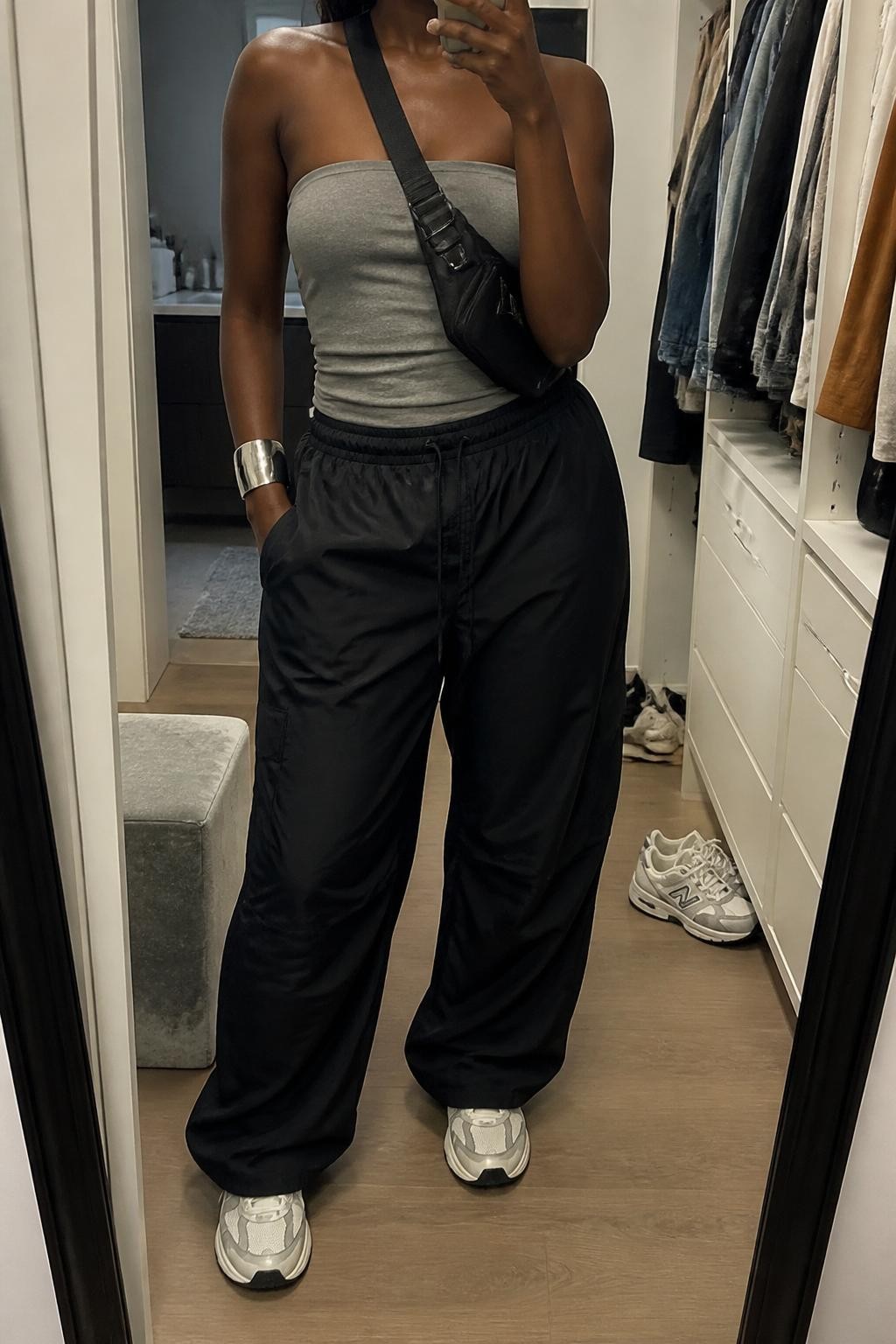 Outfit Image Prompt:
mirror selfie of a Black woman with deep brown skin wearing a gray fitted tube top, black relaxed parachute pants, white chunky sneakers, black nylon crossbody bag, and silver cuff bracelet, holding a phone in front of a mirror, cropped at NECK so the face does not show, relaxed city street style selfie pose, in a walk-in closet mirror, soft indoor lighting, with an open closet with clothes visible and a pair of sneakers on the floor nearby, realistic latest iPhone photo quality, natural skin texture.