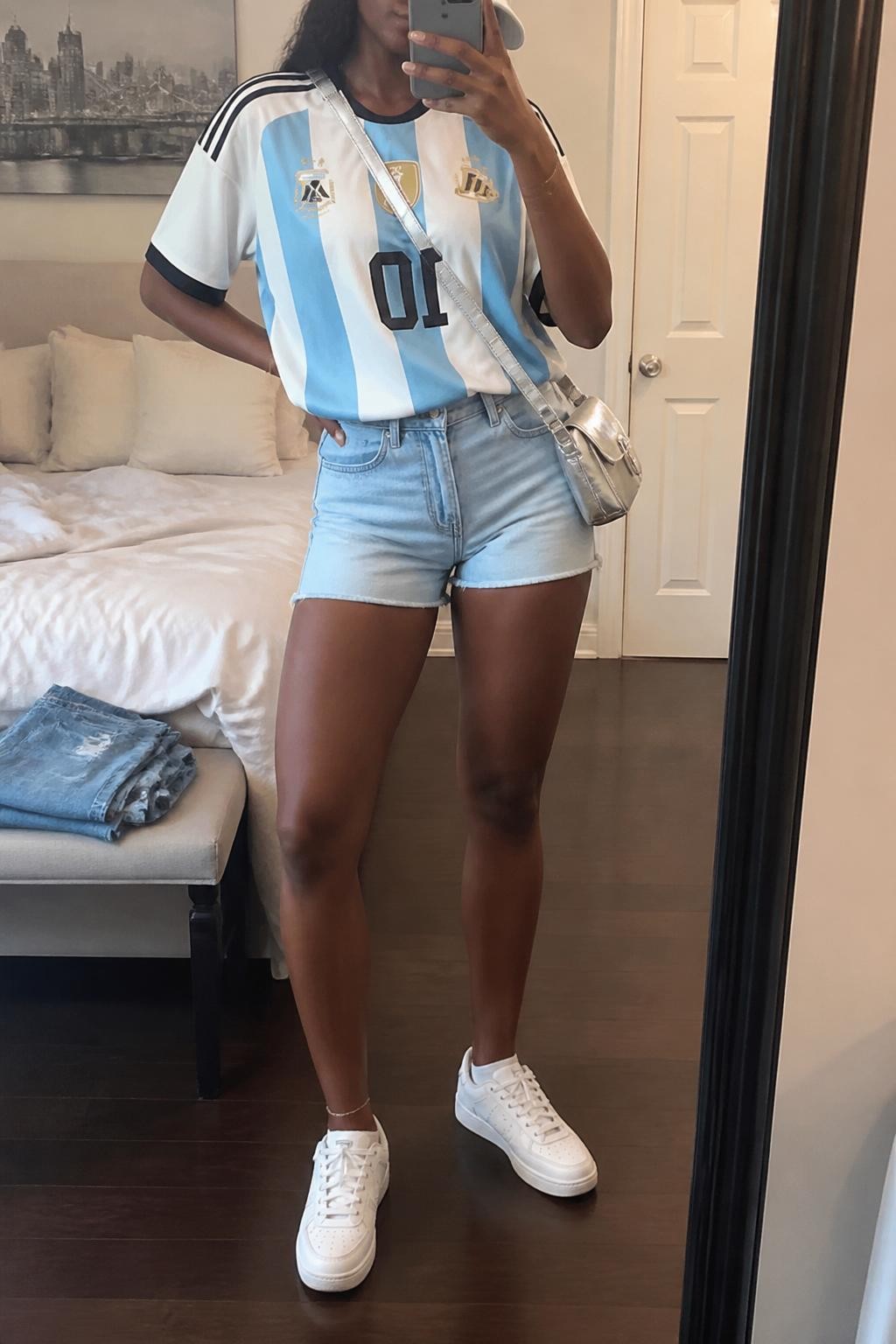 mirror selfie of a Black woman wearing an oversized sky blue and white Argentina soccer jersey, light wash high-rise denim shorts, white low-top sneakers, silver mini crossbody bag, and white baseball cap, clearly showing the same clothing items, colors, fabrics, and accessories listed in the Outfit Pieces section, holding a phone in front of a mirror, cropped at NECK so the face does not show, with a relaxed casual selfie pose, in a cozy bedroom mirror setup, soft indoor lighting, including soft pillows on an unmade bed and folded denim jeans on a bench, latest iPhone photo quality.