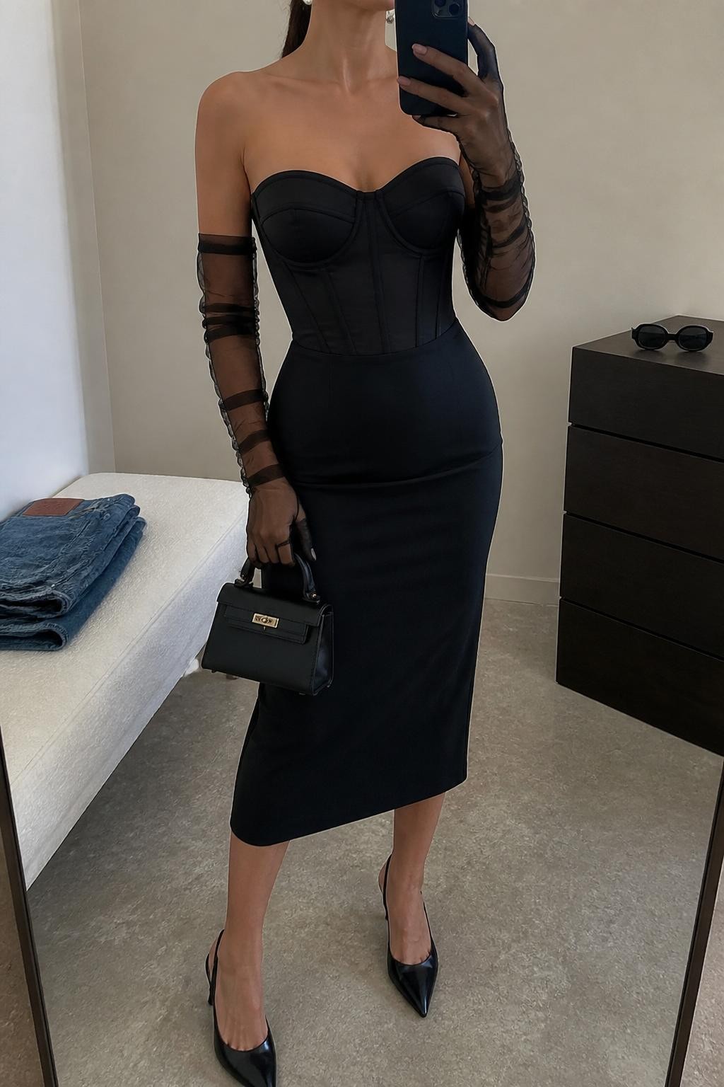 mirror selfie of a woman wearing a black corset midi cocktail dress, black mesh tulle gloves, black pointed-toe slingback heels, pearl stud earrings, a black structured mini bag, and a neat low ponytail with soft matte makeup, clearly showing the same clothing items, colors, fabrics, and accessories listed in the Outfit Pieces section, holding a phone in front of a mirror, cropped at NECK so the face does not show, with a relaxed casual selfie pose, in a minimal dressing room, soft indoor lighting, including folded denim jeans on a bench and sunglasses placed on a dresser, latest iPhone photo quality.