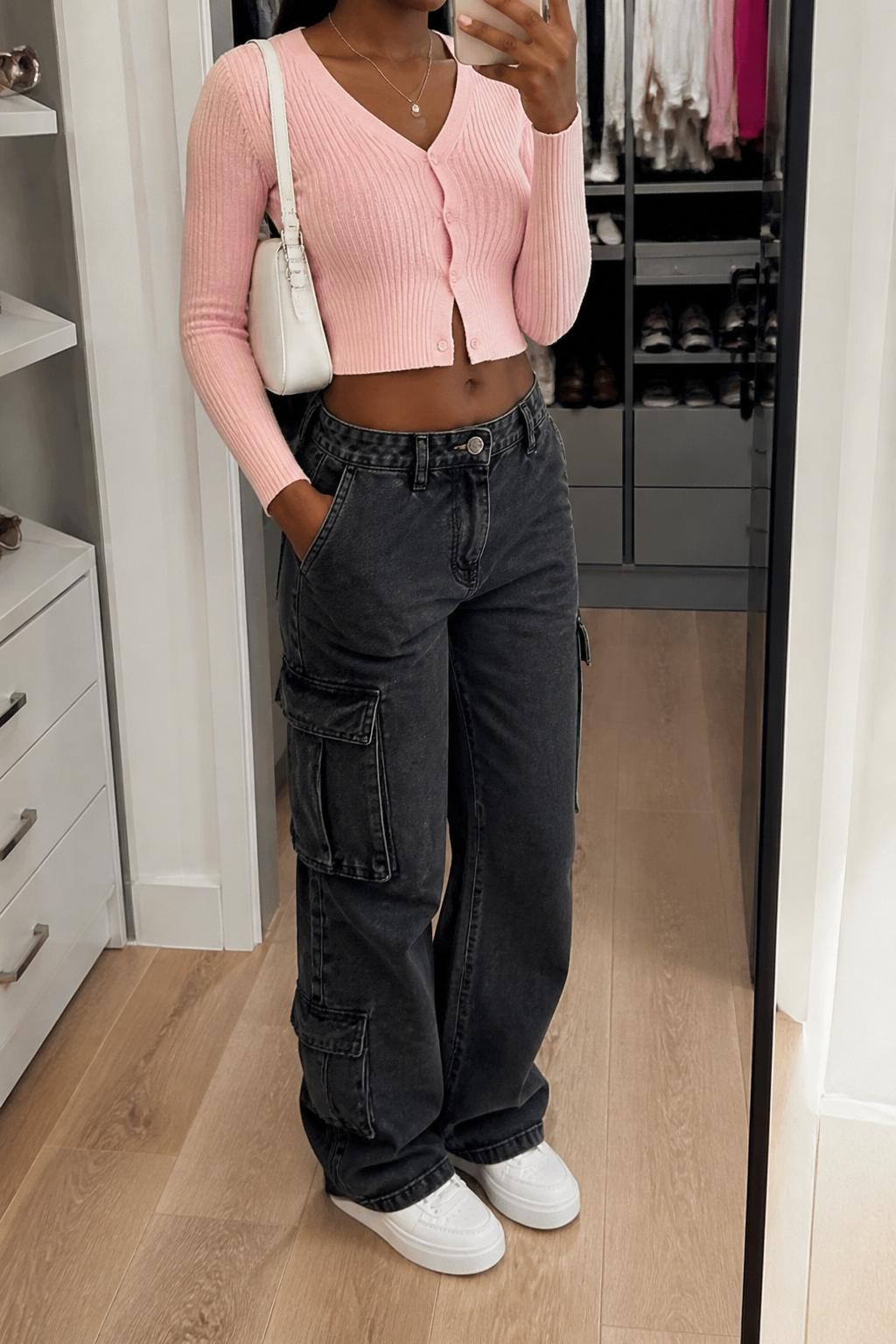 mirror selfie of a Black woman wearing washed black cargo jeans, baby pink cropped knit cardigan buttoned at the front, white platform sneakers, white mini shoulder bag, and delicate silver necklace, clearly showing the same clothing items, colors, fabrics, and accessories listed in the Outfit Pieces section, holding a phone in front of a mirror, cropped at NECK so the face does not show, with a relaxed casual selfie pose, in a walk-in closet mirror, soft indoor lighting, including an open closet with clothes visible and sunglasses placed on a dresser, latest iPhone photo quality.