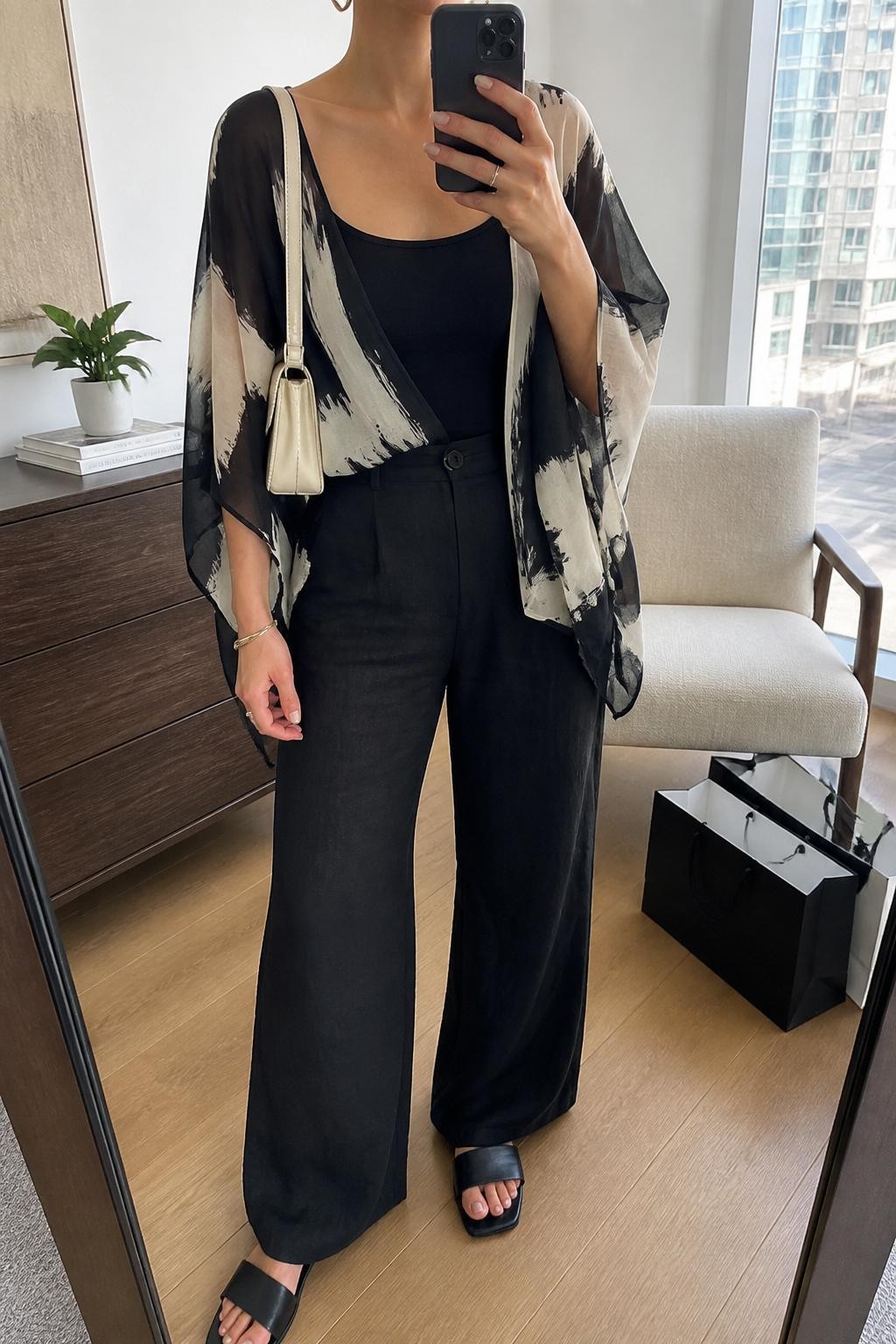 mirror selfie of a woman wearing a black and ivory abstract printed sheer poncho layered over a black fitted camisole, black high-waisted linen pants, black leather slide sandals, ivory mini shoulder bag, and gold hoop earrings, clearly showing the same clothing items, colors, fabrics, and accessories listed in the Outfit Pieces section, holding a phone in front of a mirror, cropped at NECK so the face does not show, with a relaxed casual selfie pose, in a stylish city apartment, natural window light, including shopping bags beside a chair and a small plant on a dresser, latest iPhone photo quality.