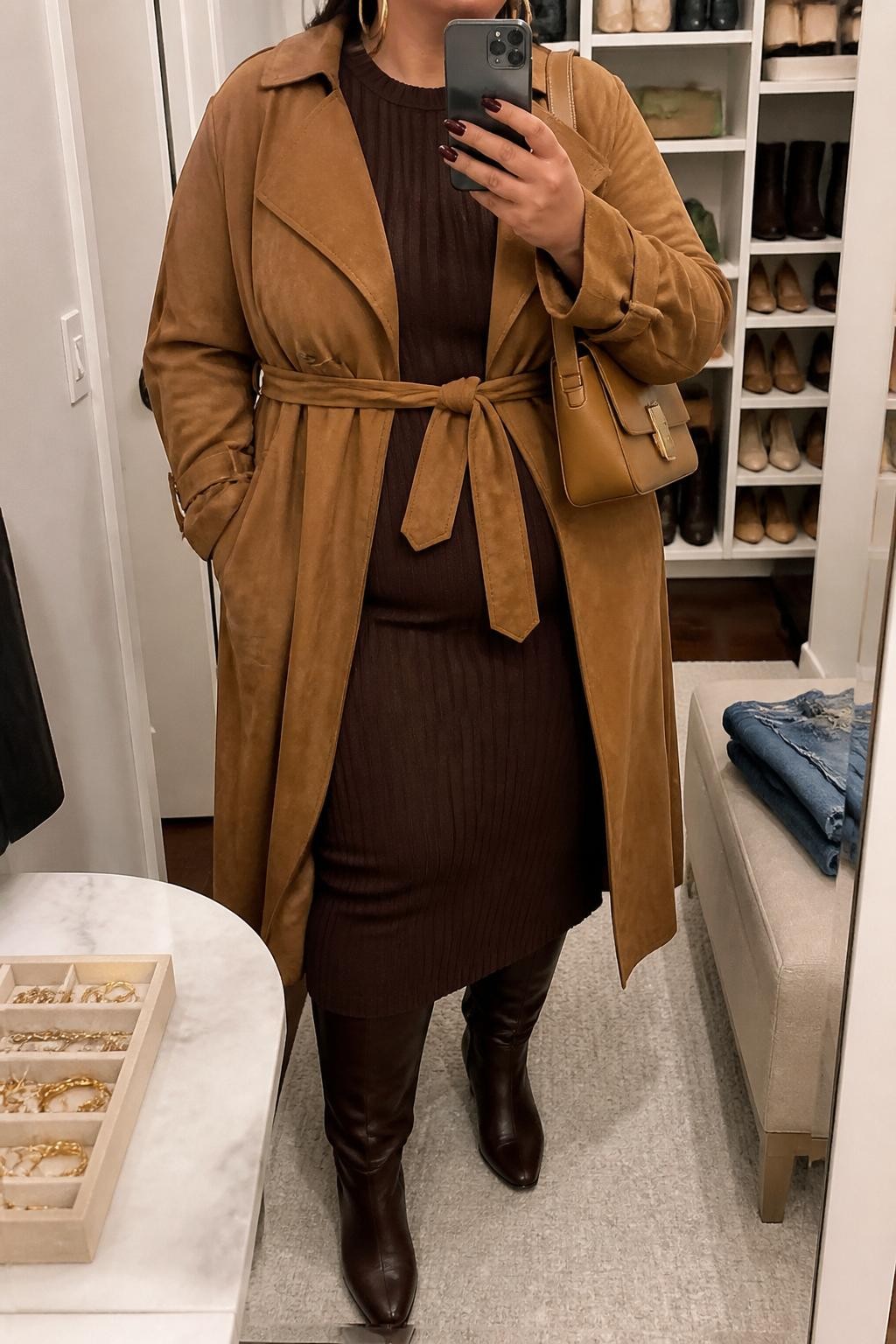 mirror selfie of a plus size woman wearing the exact outfit described above, a warm camel suede trench coat with a waist tie layered over a rich chocolate brown ribbed knit midi dress, dark brown wide-calf knee-high boots, tan structured shoulder bag, and gold hoop earrings, clearly showing the same clothing items, colors, fabrics, and accessories listed in the Outfit Pieces section, holding a phone in front of a mirror, cropped at NECK so the face does not show, with a relaxed casual selfie pose, in a walk-in closet mirror, soft indoor lighting, including a jewelry tray on the vanity and folded denim jeans on a bench, latest iPhone photo quality.